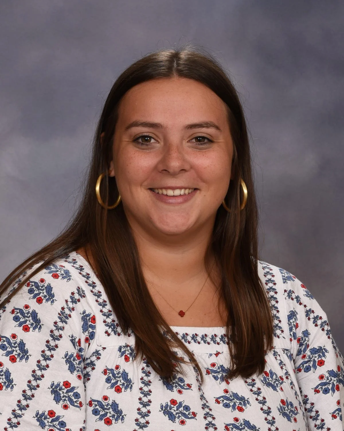 A young woman with brown hair, wearing large gold hoop earrings, a white blouse with blue and red floral embroidery, and a small red flower necklace, smiling against a gray background.
