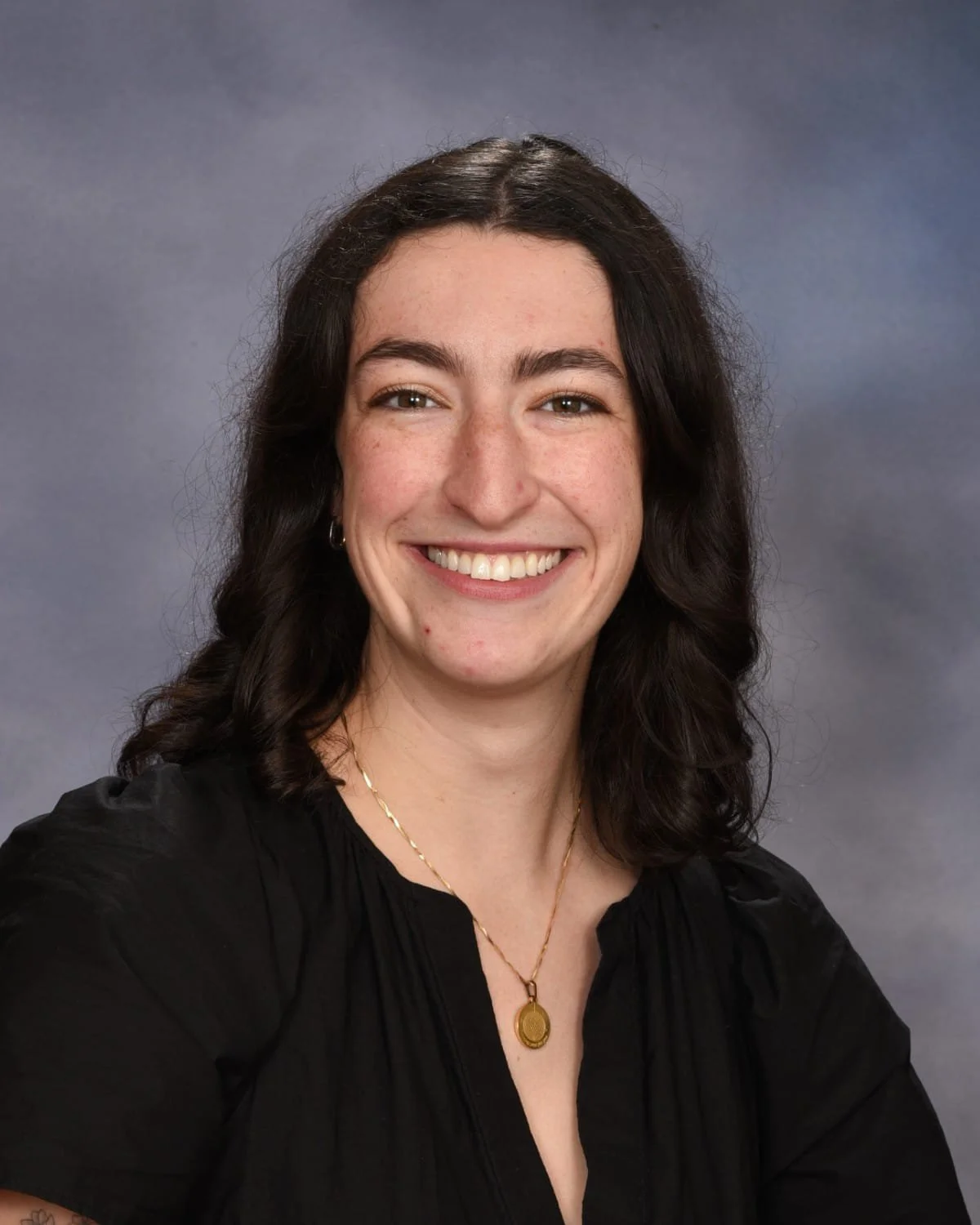 A woman with dark wavy hair, smiling, wearing a black top, a gold necklace with a round pendant, and small hoop earrings. She has a light complexion and is posing in front of a gray background.