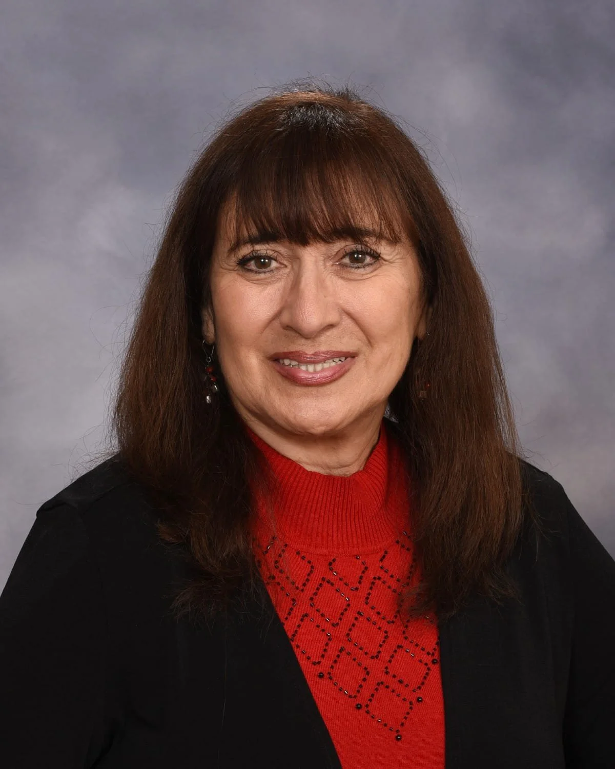 A woman with long brown hair, wearing a red sweater with black diamond patterns and a black blazer, smiling against a neutral background.