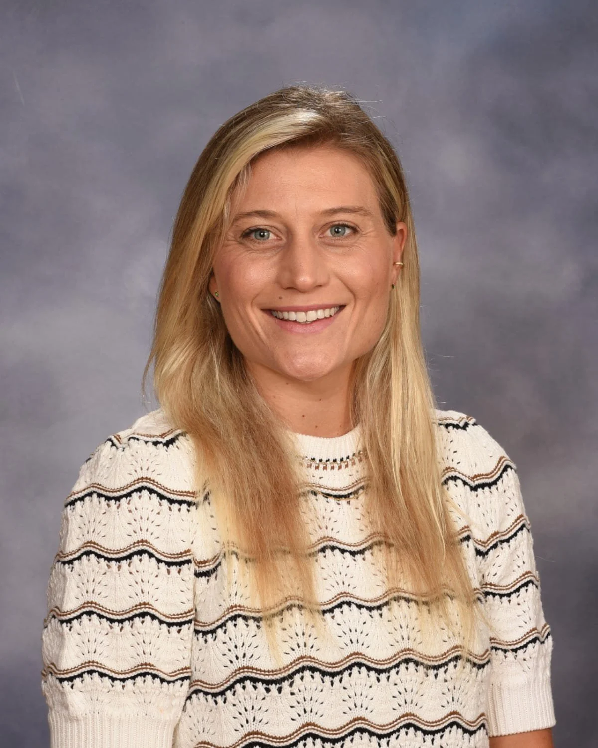 A smiling blonde woman wearing a cream and black patterned sweater against a gray background.