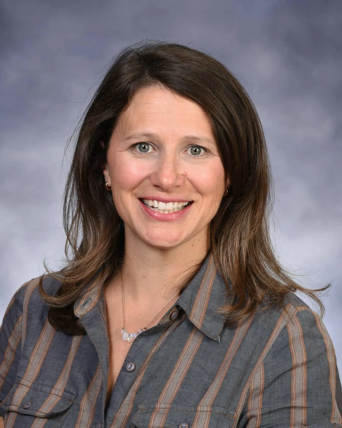 A woman with shoulder-length brown hair, wearing a grey button-up shirt with brown stripes, smiling against a cloudy grey background.