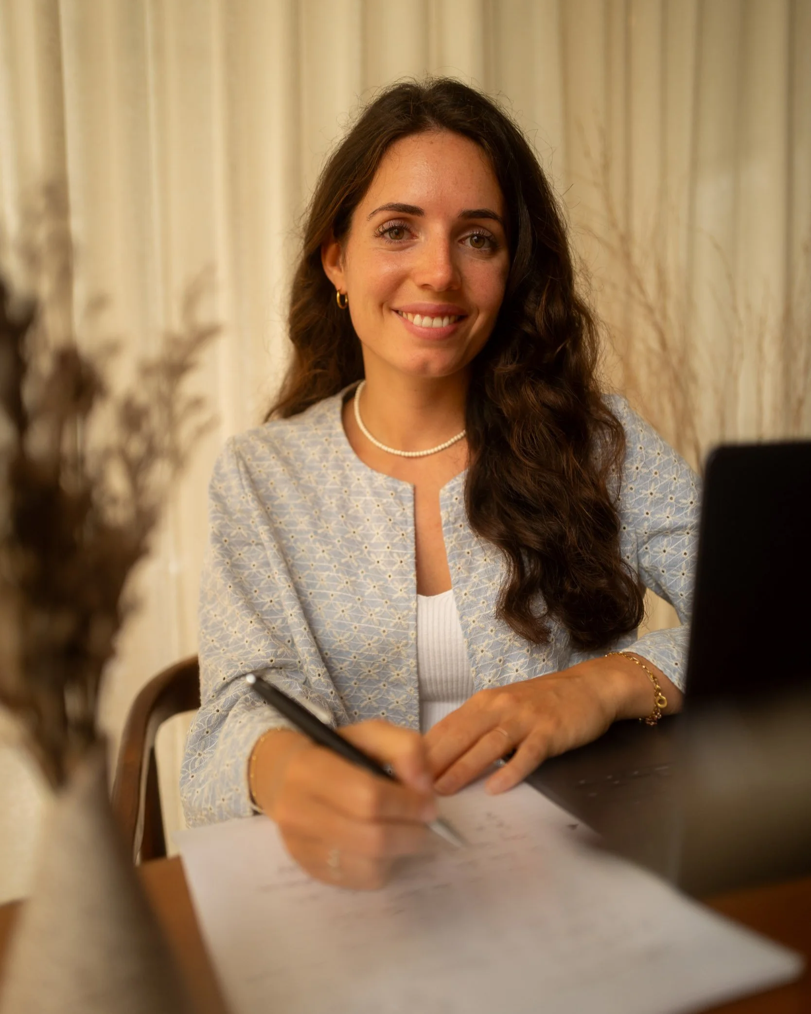 Eine lächelnde Frau mit braunen, lockigen Haaren, die an einem Tisch sitzt, während sie schreibt und einen Laptop vor sich hat, vor einem Vorhang.