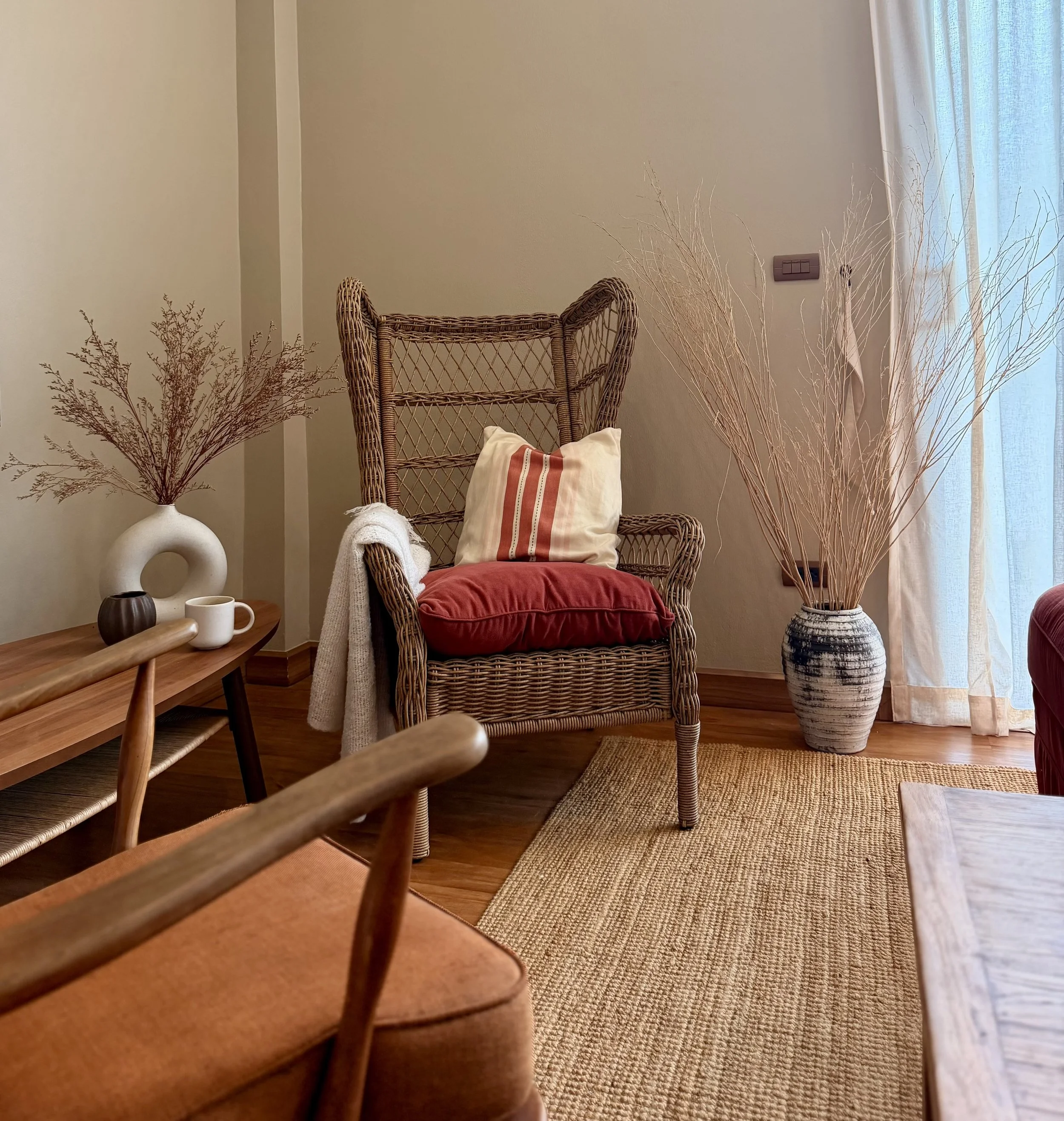 Ein Wohnzimmer mit einem Rattanstuhl, dekorativen Kissen, einem großen Vase mit Zweigen, einem Couchtisch, einem Teppich und Vorhängen.