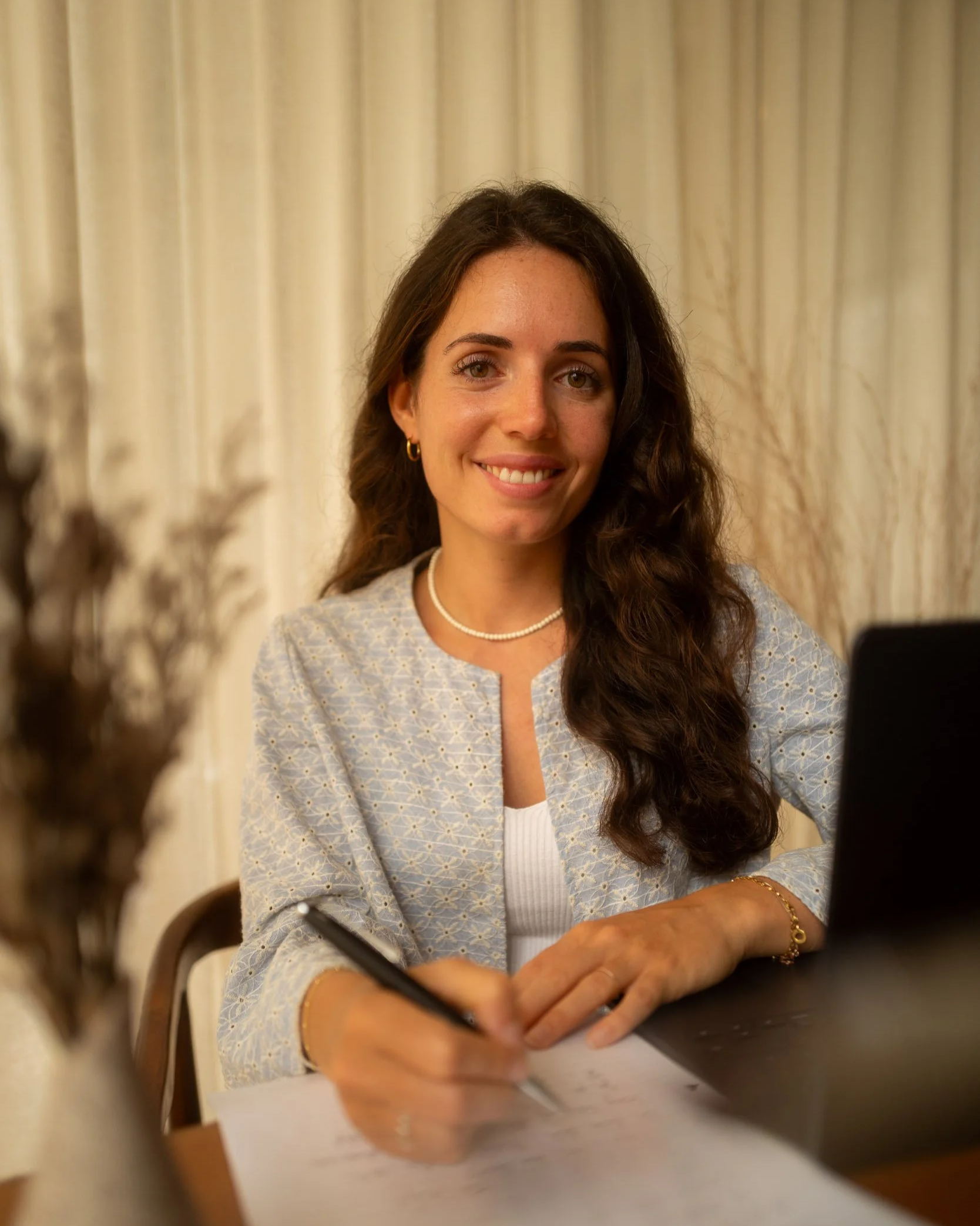 Eine lächelnde Frau sitzt an einem Tisch mit einem Notizbuch und einem Laptop, im Hintergrund ein Vorhang.