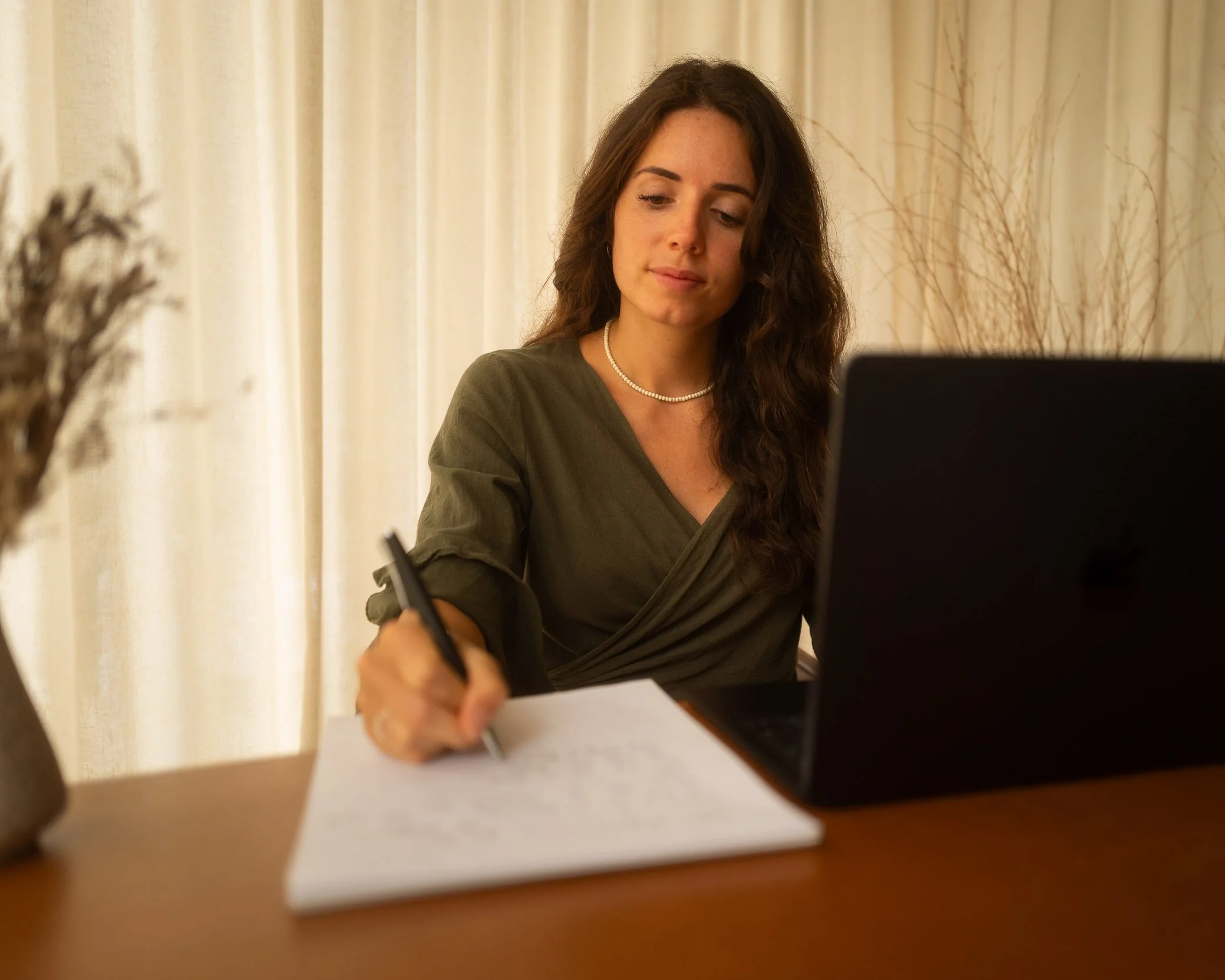 Frau arbeitet an einem Schreibtisch mit Laptop und Notizbuch in einem Raum mit weißen Vorhängen