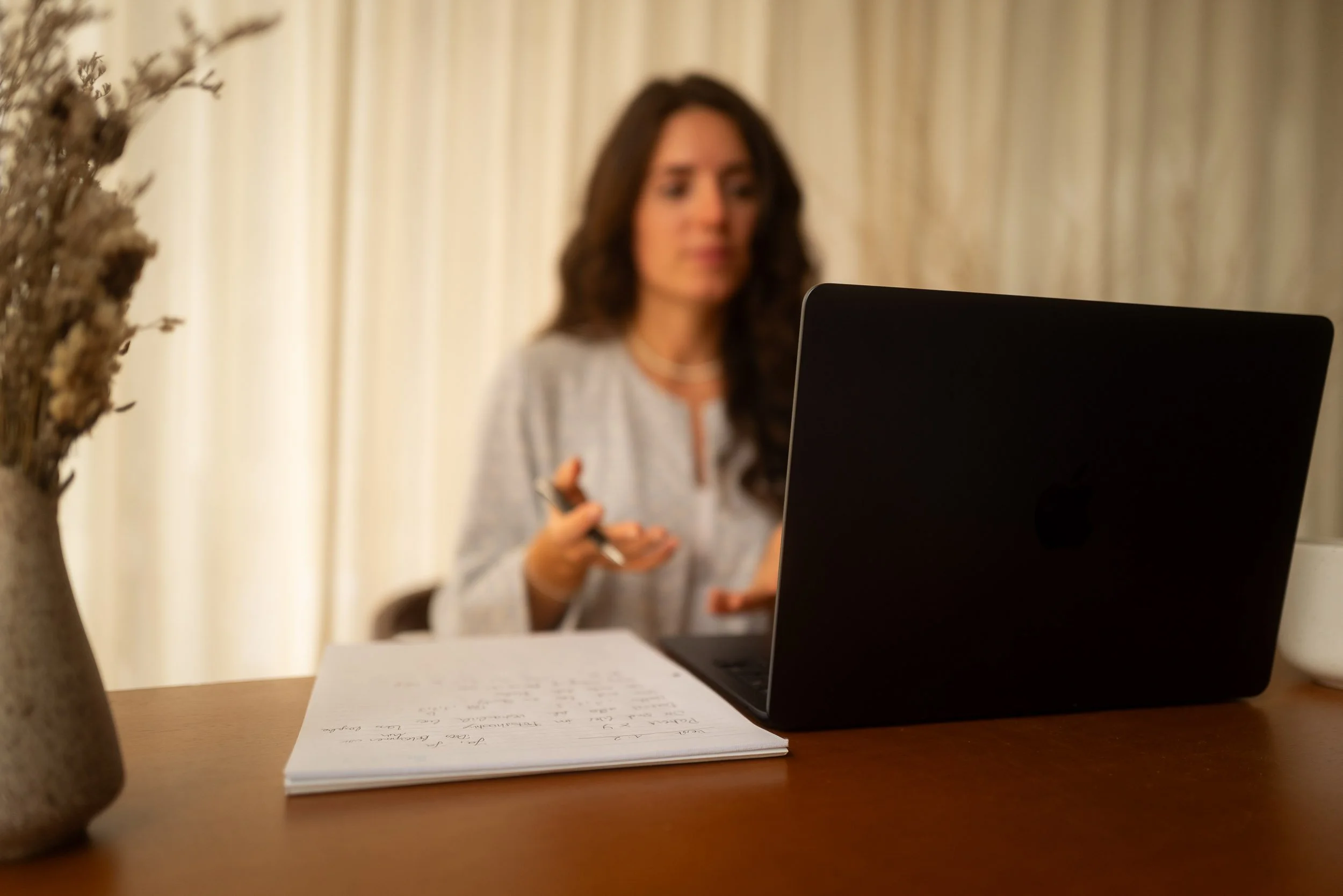 Eine Frau sitzt vor einem Laptop, im Vordergrund liegen Notizen auf einem Tisch, und eine Vase mit getrockneten Blumen ist sichtbar.