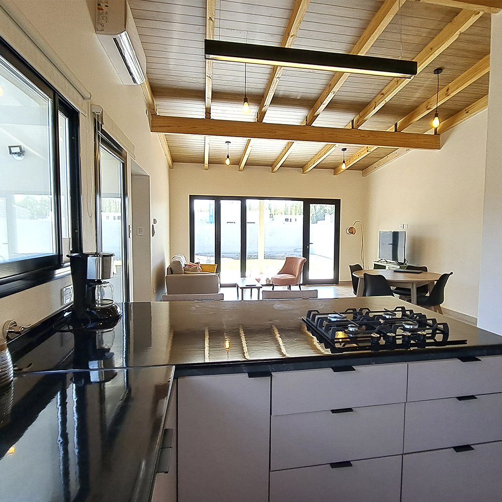 Open-concept living space with a kitchen island in the foreground, and a living room with a beige sofa, pink armchair, and a small round table in front of large sliding glass doors leading outside. There's a dining area nearby with a round table and