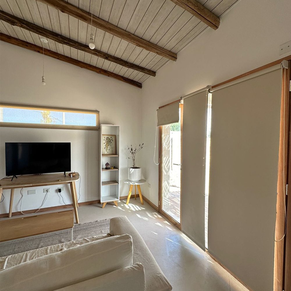 Living room with a TV on a wooden stand, a white bookshelf, a potted plant on a small yellow table, a beige sofa, large window and glass door with blinds, and a vaulted wooden ceiling.