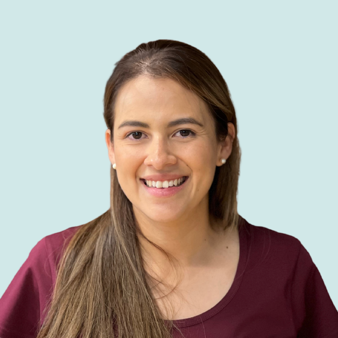 A woman with long brown hair, wearing pearl earrings and a maroon T-shirt with a logo, smiling against a light purple background.