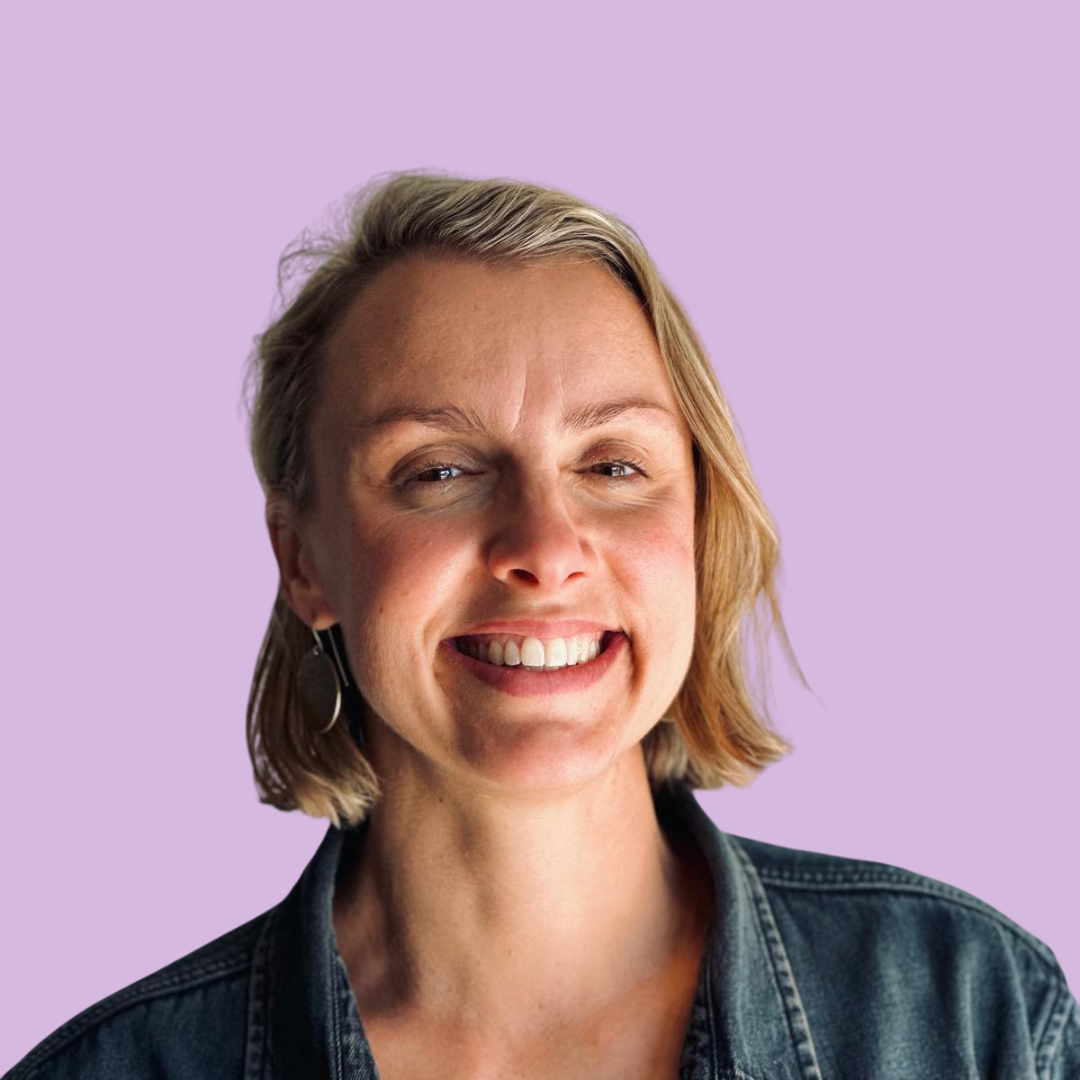 Close-up portrait of a woman with blonde hair, smiling, wearing earrings and a denim jacket against a light blue background.