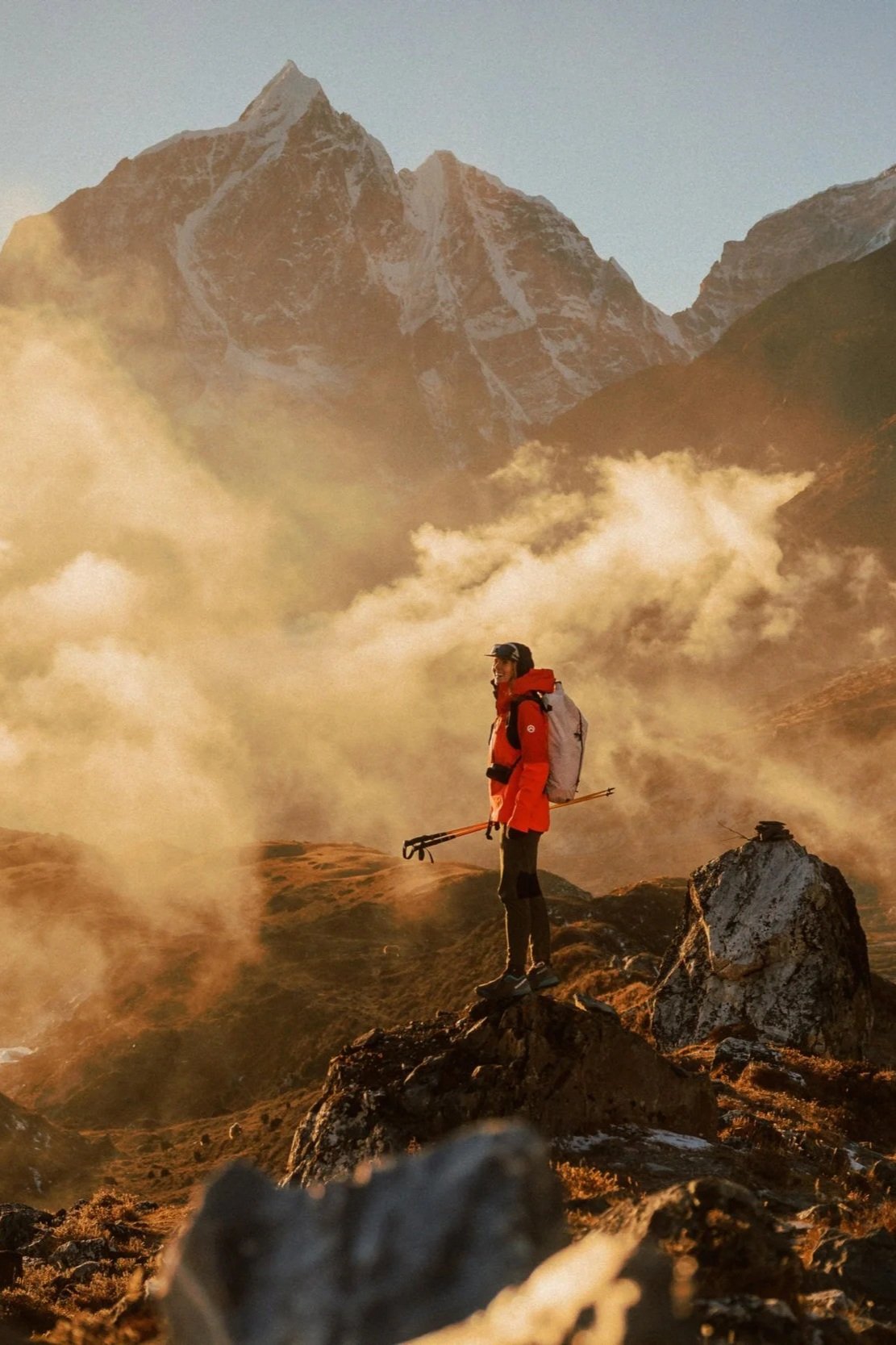 Ein Bergsteiger in roter Jacke steht auf einem Felsen in einer bergigen Landschaft mit Wolken und hohen Gipfeln im Hintergrund, bei Sonnenaufgang oder Sonnenuntergang.