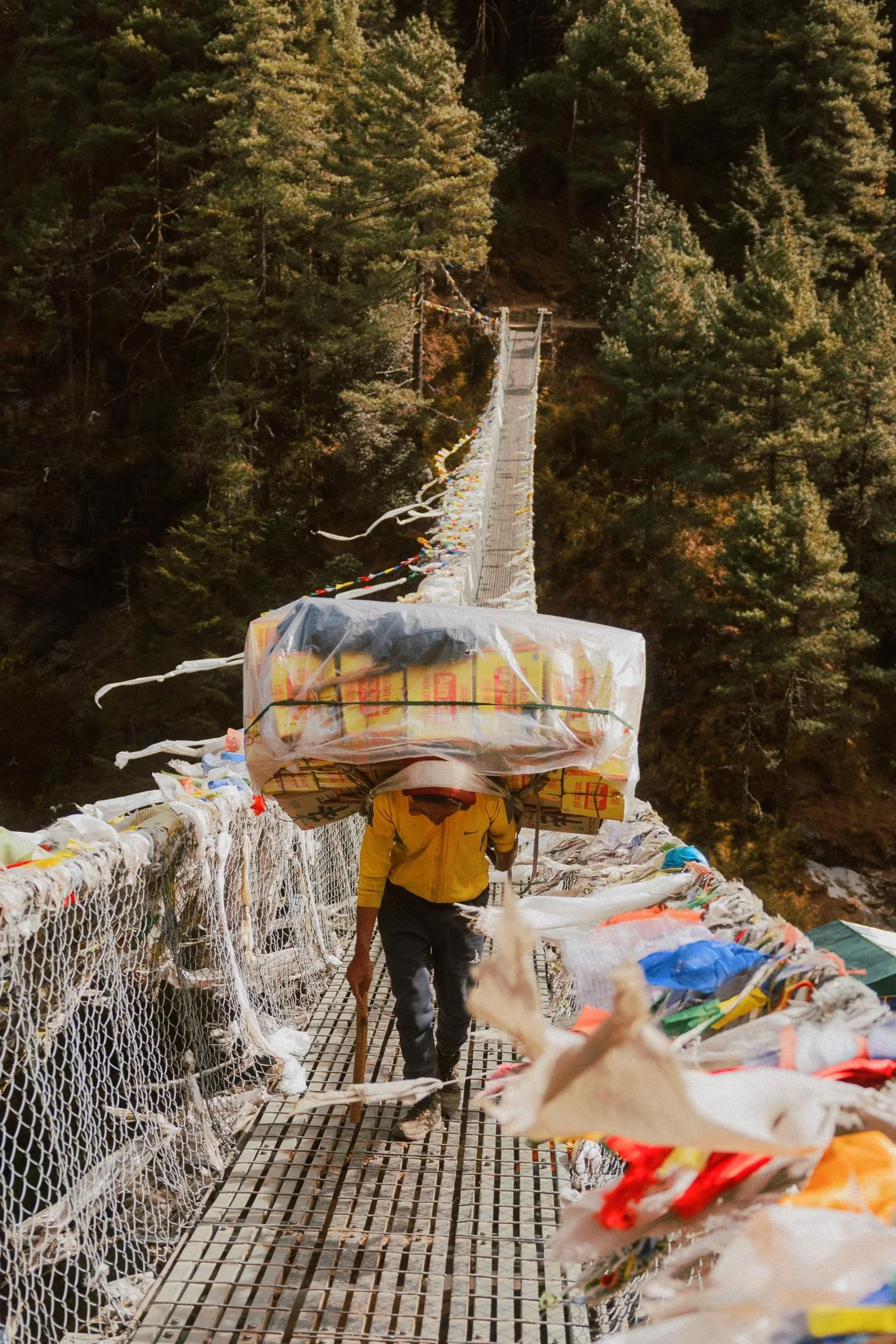 Ein Mann trägt schwere Pakete über eine schmale Hängebrücke in einem Wald. Die Brücke ist mit bunten Bändern und Plastikfolien dekoriert.