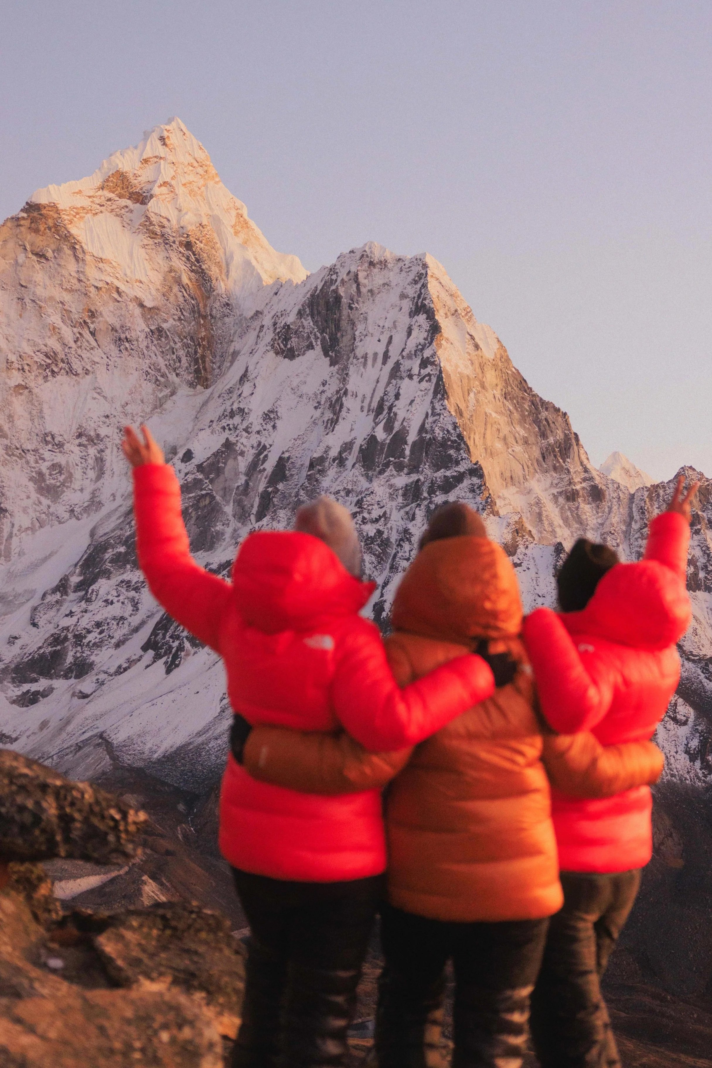 Drei Menschen in Daunenjacken stehen mit dem Rücken zur Kamera, schauen auf schneebedeckte Berge bei Sonnenaufgang und heben die Arme zum Himmel. Ein Gefühl von Freiheit, Lebensfreude und Abenteuer auf einer Gruppenreise mit Aspire Adventures erleben