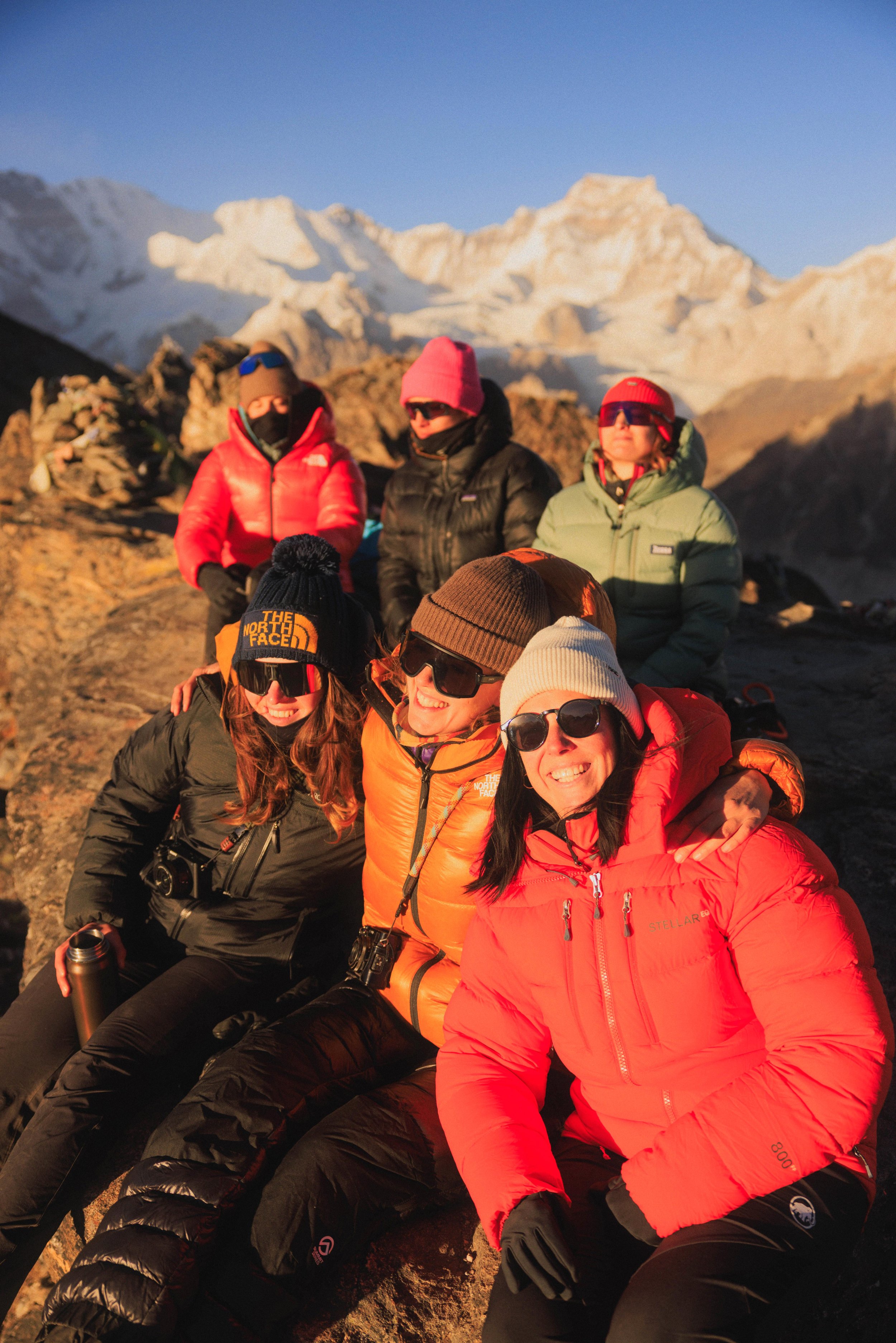 Sechs Personen in Winterkleidung auf einem Berggipfel mit Schnee und Bergen im Hintergrund, die Sonne scheint.