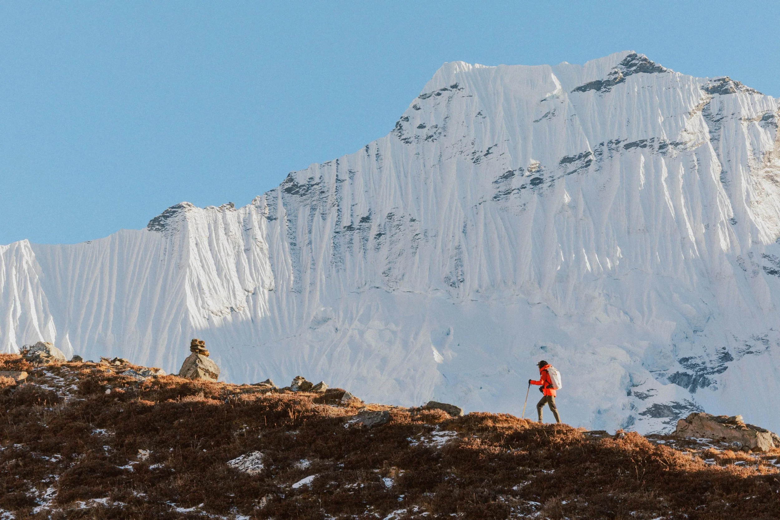 Ein Wanderer in rot und grau auf einem Berghang mit Gestein und Büschen, im Hintergrund eine große schneebedeckte Gebirgsklippe. Wandern in Nepal. Trekking zum Everest Basecamp.  Chukhung. Gruppenreise Nepal. Fotografie Reise.