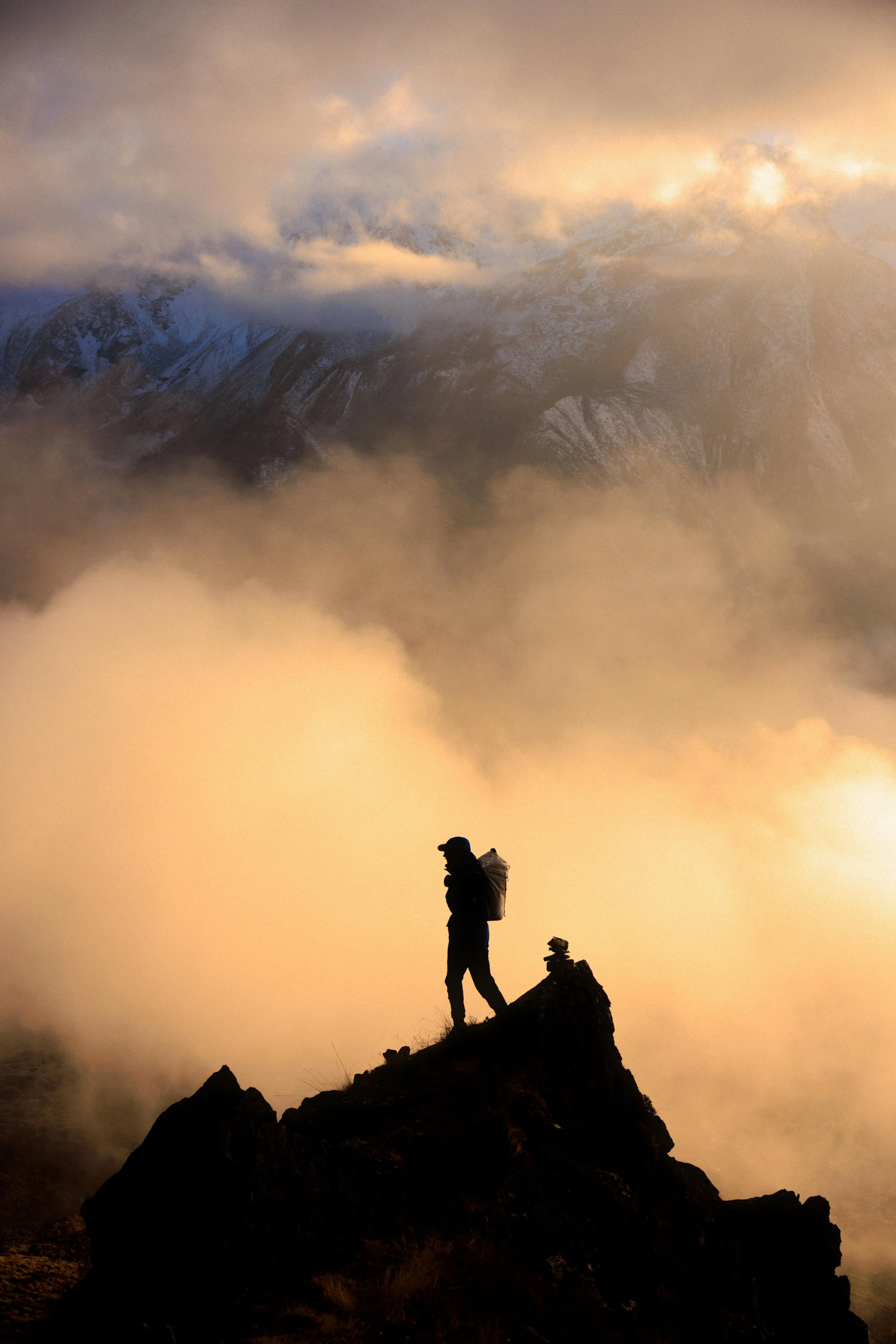 Ein einsamer Bergsteiger mit Rucksack auf einem felsigen Gipfel bei Sonnenaufgang, umgeben von Wolken und Nebel, mit verschneiten Bergen im Hintergrund.