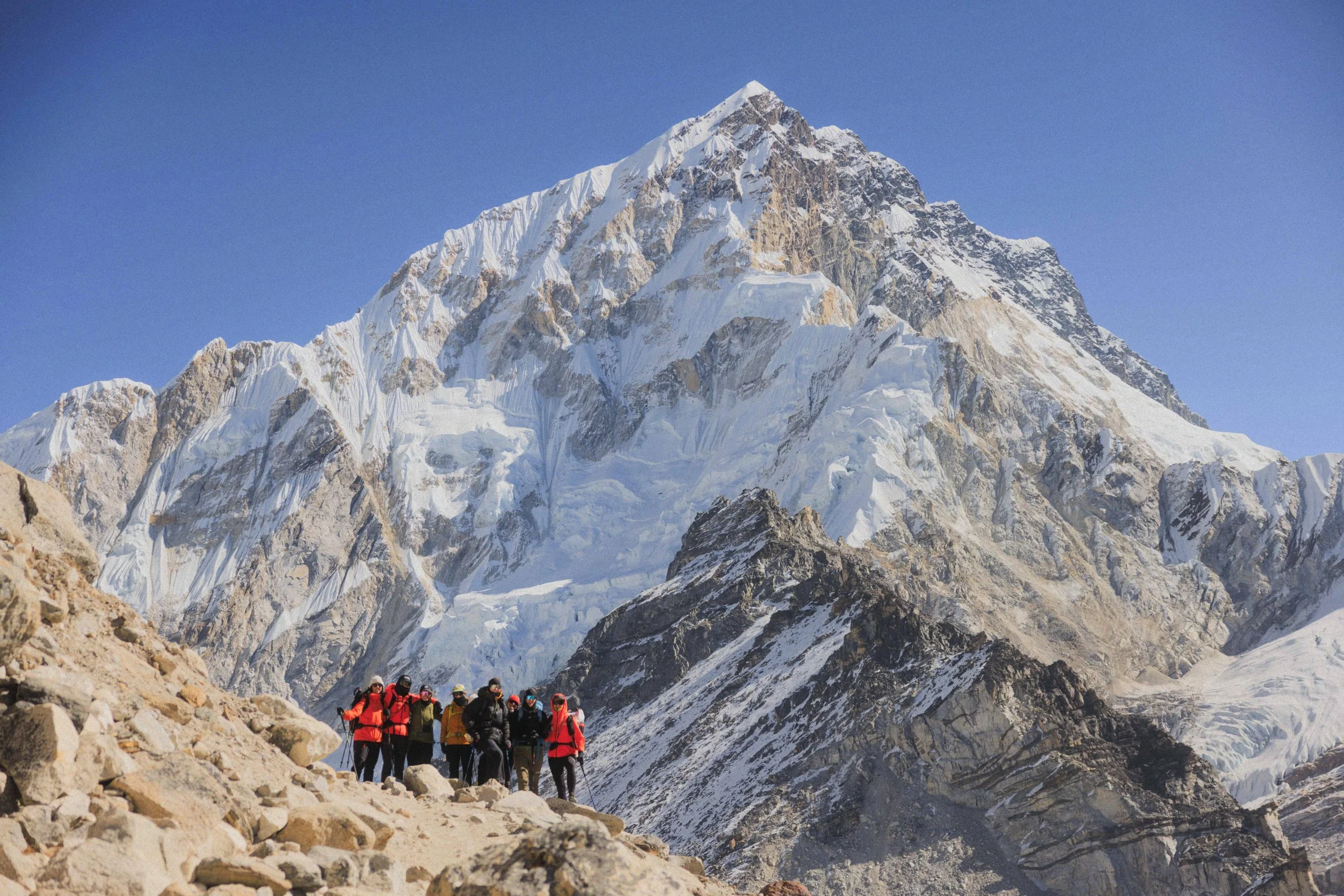 Eine Gruppe von Wanderern in bunter Kleidung auf einem felsigen Weg vor einer großen, schneebedeckten Bergkette mit Gipfel unter blauem Himmel.