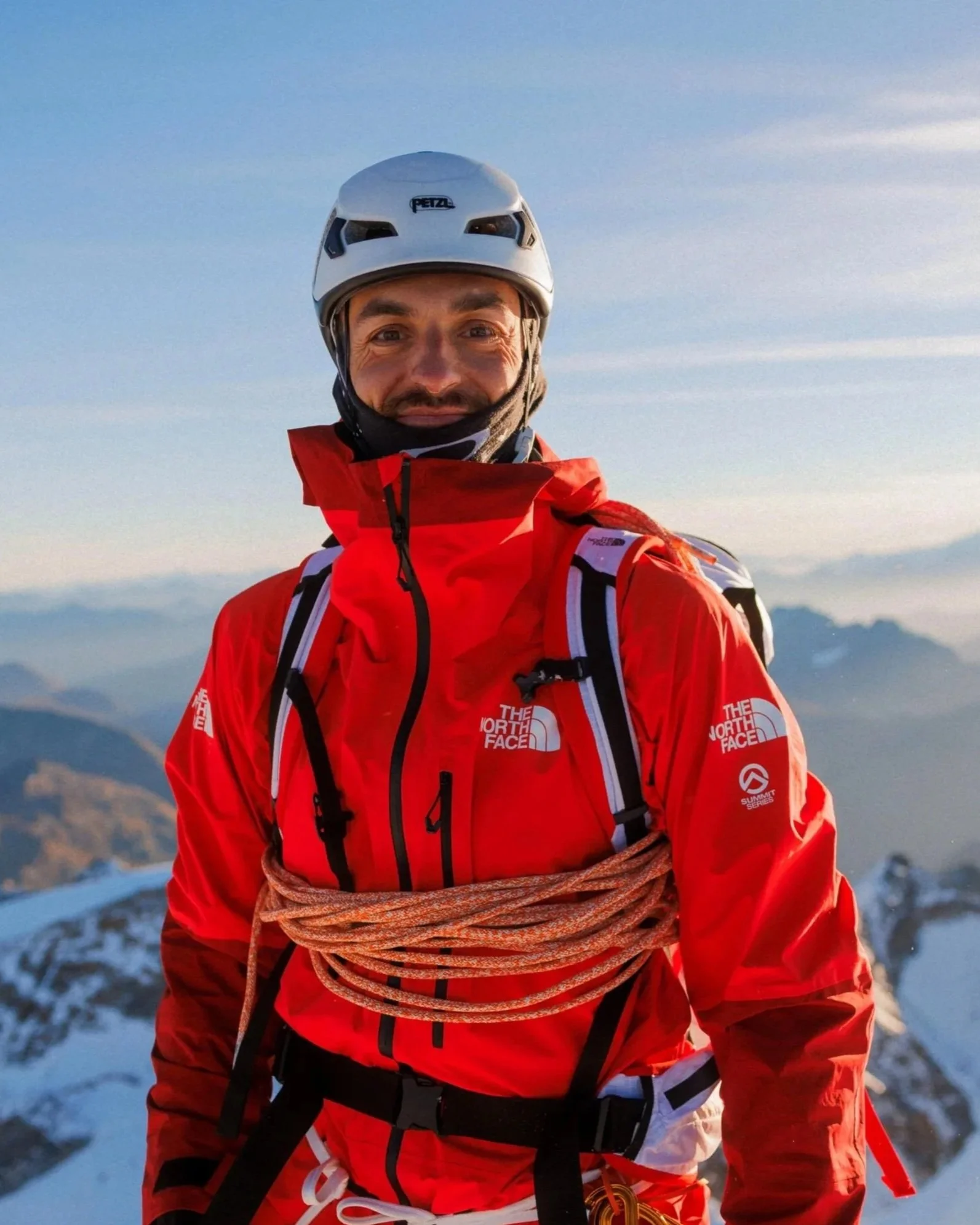Bergsteiger trägt rote Jacke, Klettersicherung um den Oberkörper, Helm und Rucksack vor einer Bergkulisse mit Schnee. David Herzig. Reiseleitung. Nepal Reisen. Gruppenreise Nepal. Bergfotografie. Fotografie Kurs.