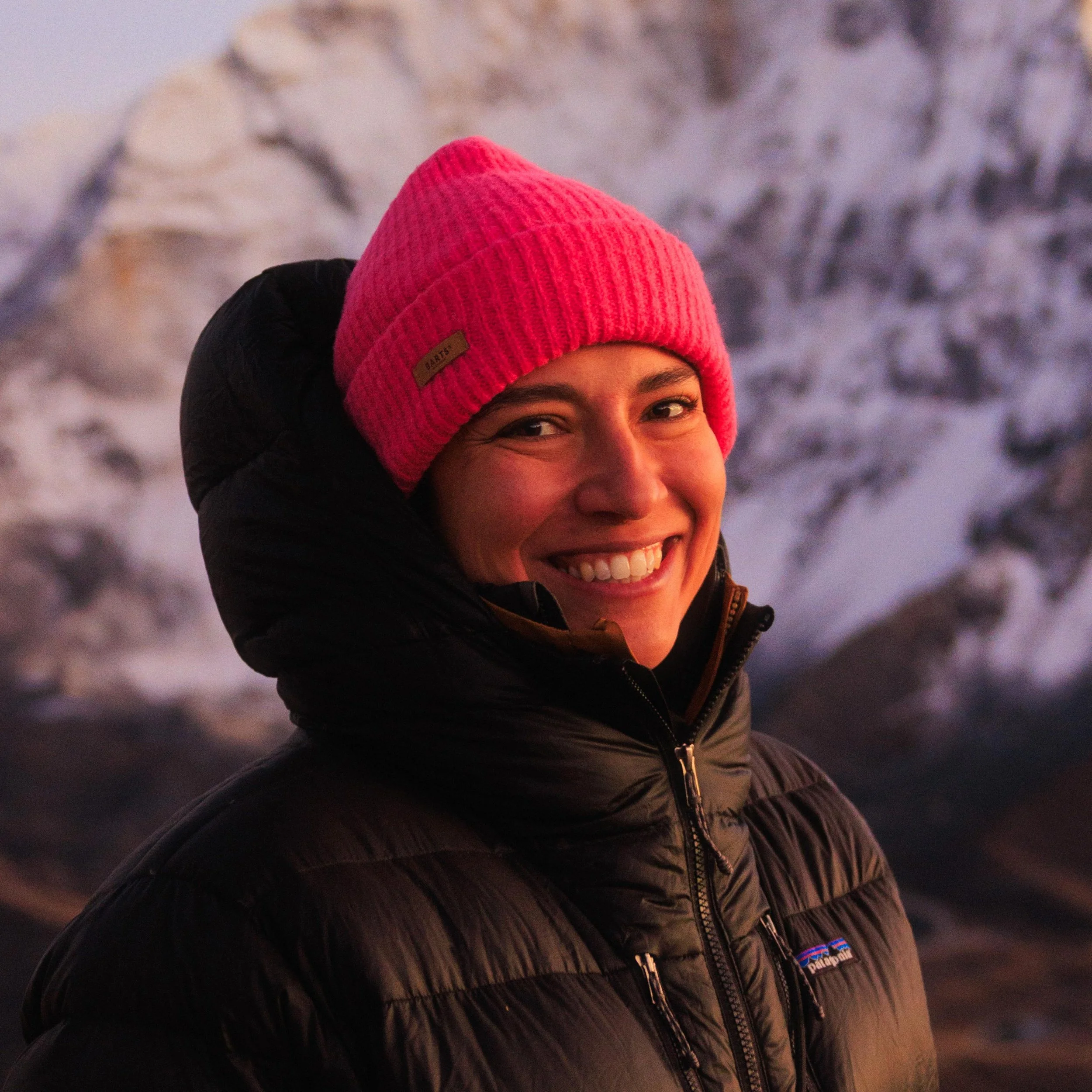 Lächelnde Frau mit pinker Mütze und schwarzer Winterjacke vor einer verschneiten Bergkulisse bei Sonnenuntergang.