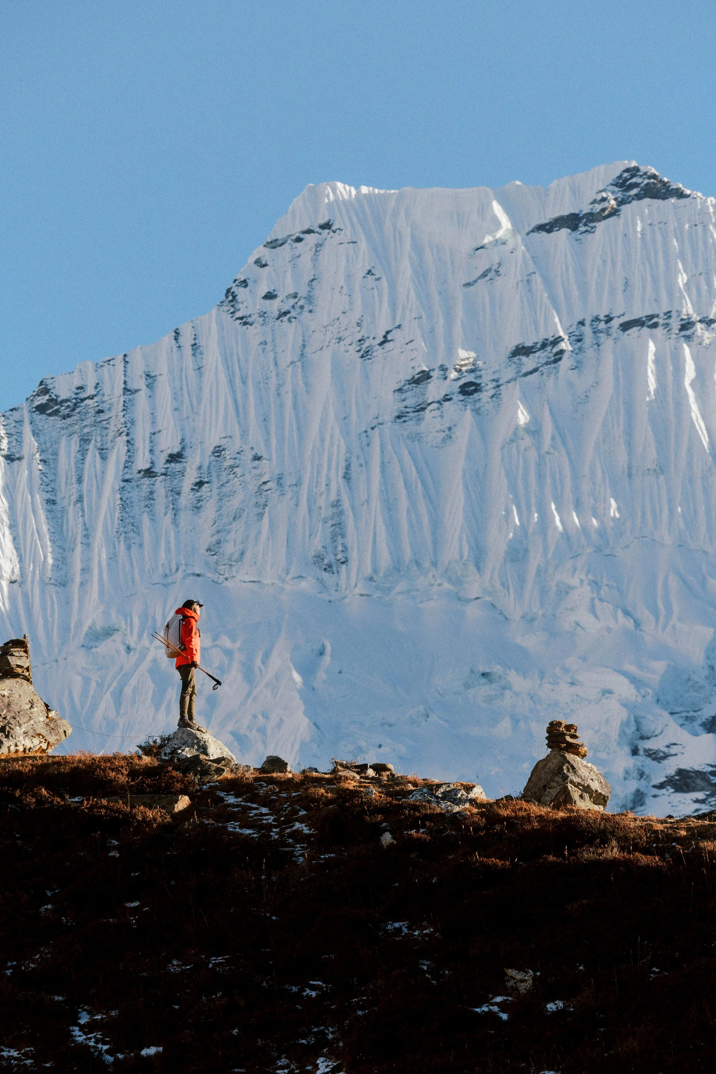 Eine Person in rot-warmer Kleidung mit Skihandschuhen steht auf einem Felsen vor einer riesigen, schneebedeckten Gletscherwand, während die Sonne das Eis beleuchtet.