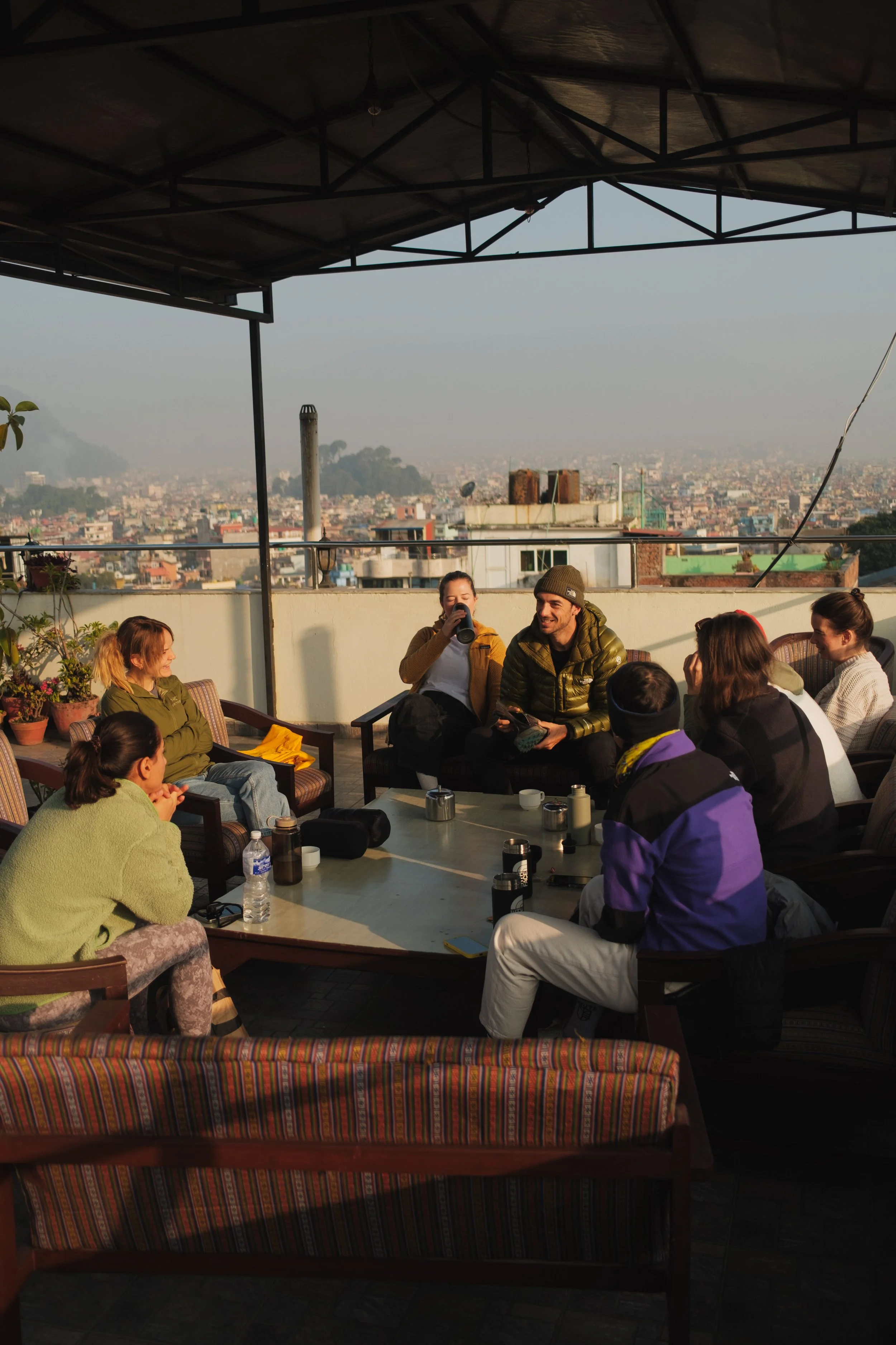 Menschen sitzen auf einer Dachterrasse und unterhalten sich bei Sonnenuntergang, mit Stadtblick im Hintergrund.