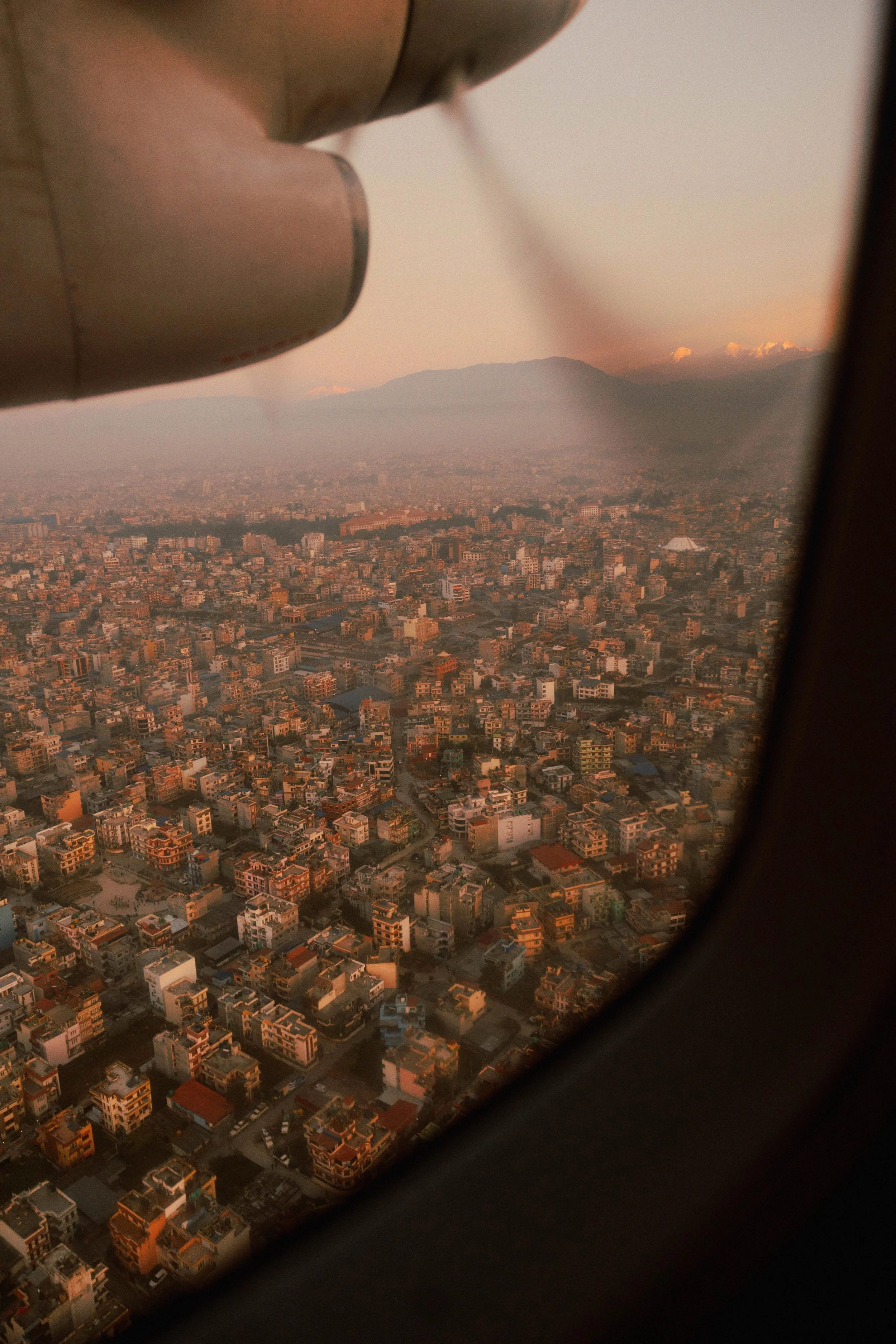 Luftaufnahme einer Stadt bei Sonnenuntergang, aufgenommen durch das Flugzeugfenster, mit einer Propellermaschine im Vordergrund.