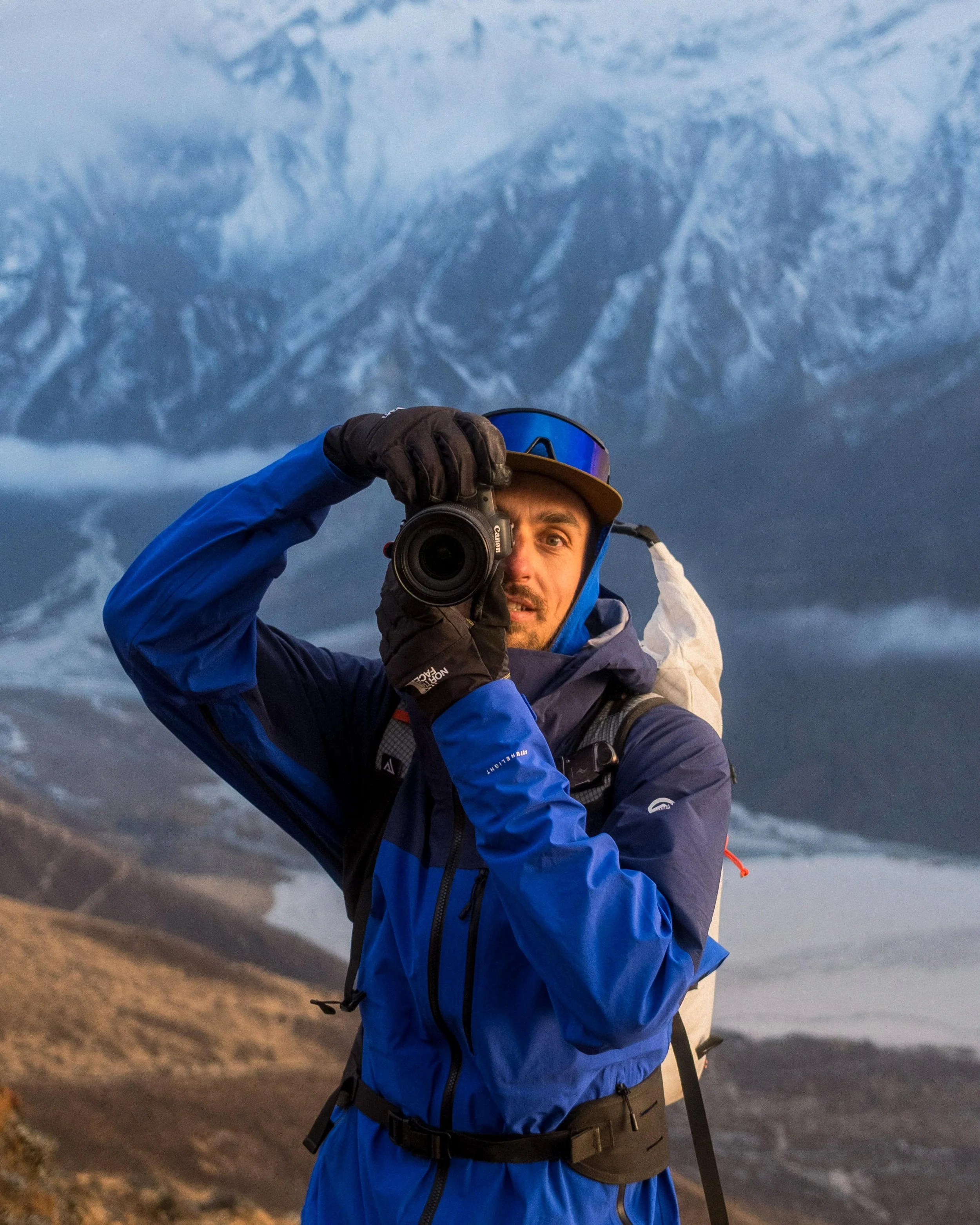 Ein Mann in Outdoor-Bekleidung nimmt ein Selfie mit einer Kamera vor einer Berglandschaft mit Gletschern und Bergen auf.
