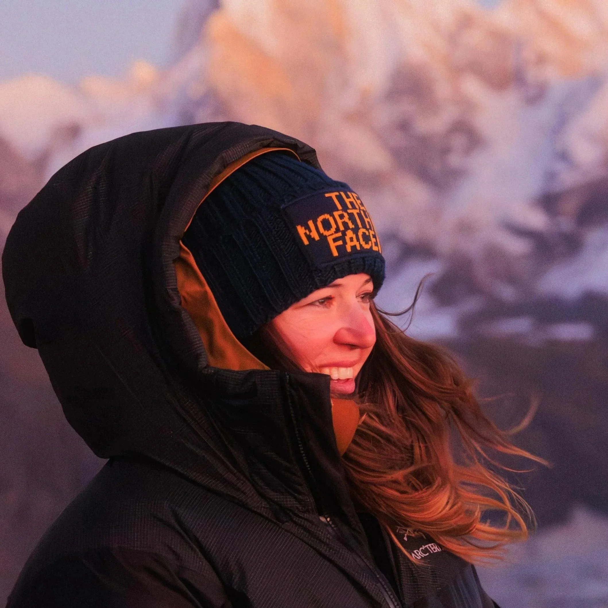 Junge Frau mit Wind im Haar, trägt eine schwarze Jacke und eine Mütze mit dem Logo 'The North Face', auf einem Berggipfel bei Sonnenuntergang.