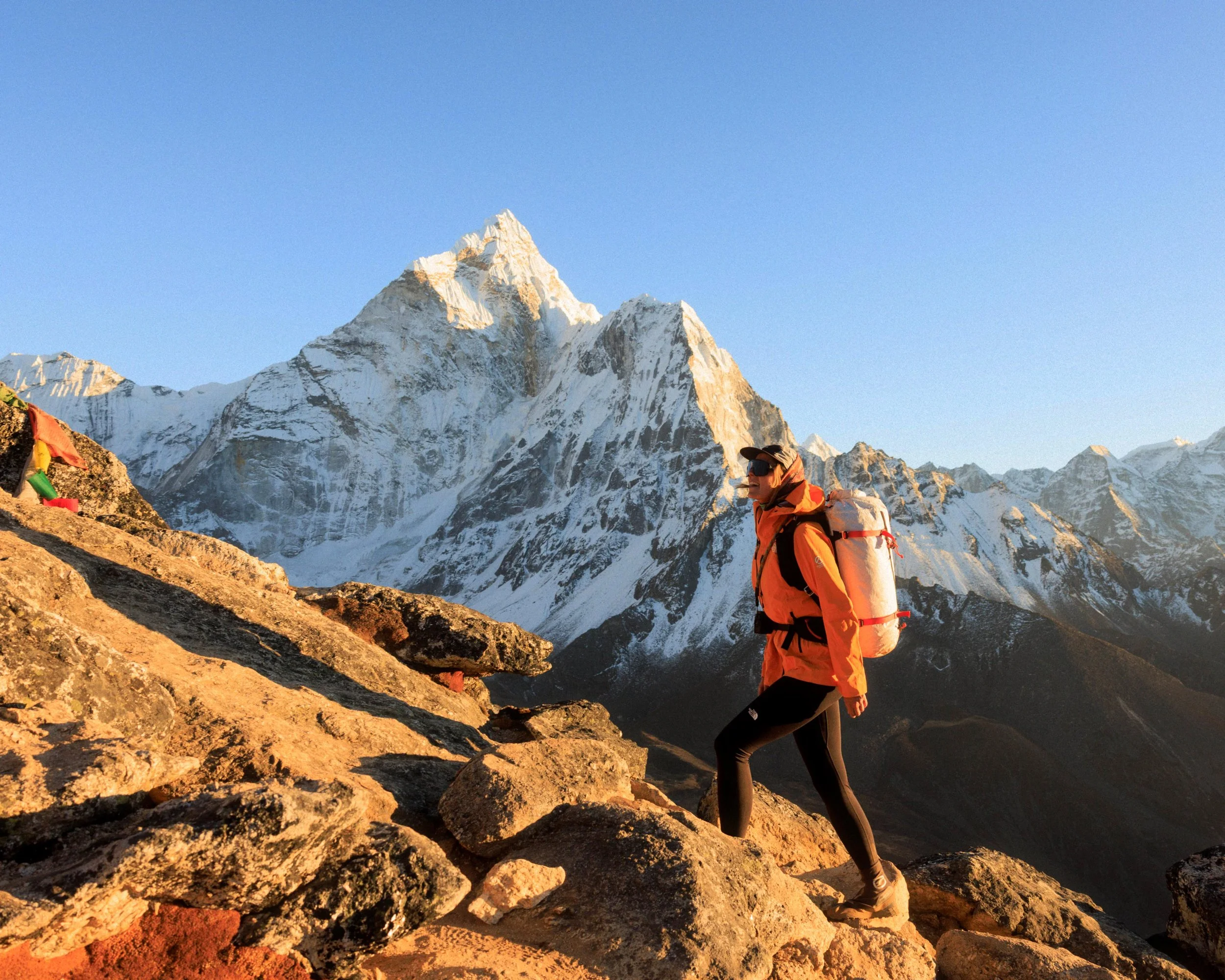 Eine Bergsteigerin beim Klettern in einer Gebirgslandschaft mit schneebedeckten Bergen im Hintergrund, Sonnenlicht scheint auf die Szene.