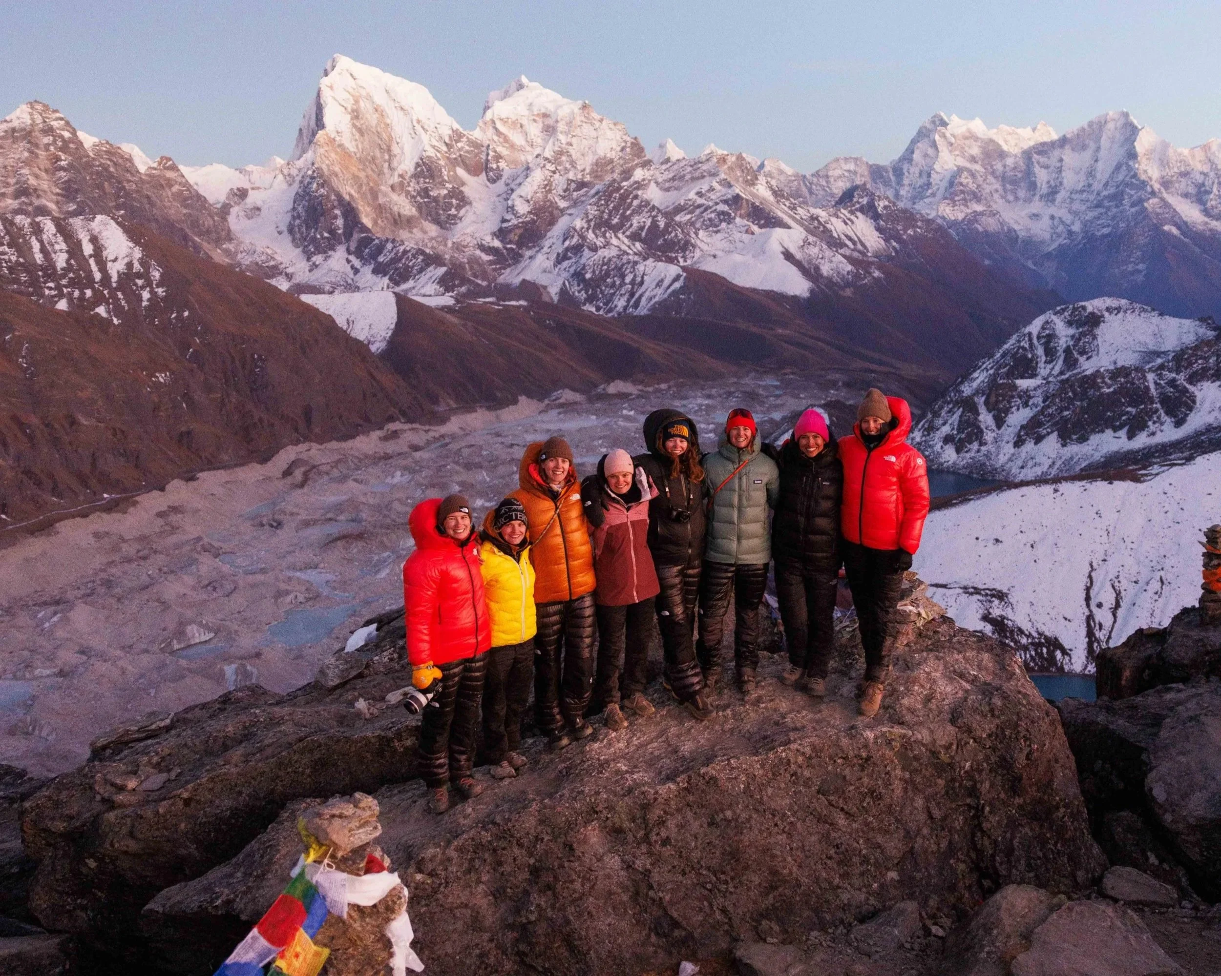 Sieben Menschen in warmer Kleidung auf einem Felsen in den Bergen mit schneebedeckten Gipfeln im Hintergrund.