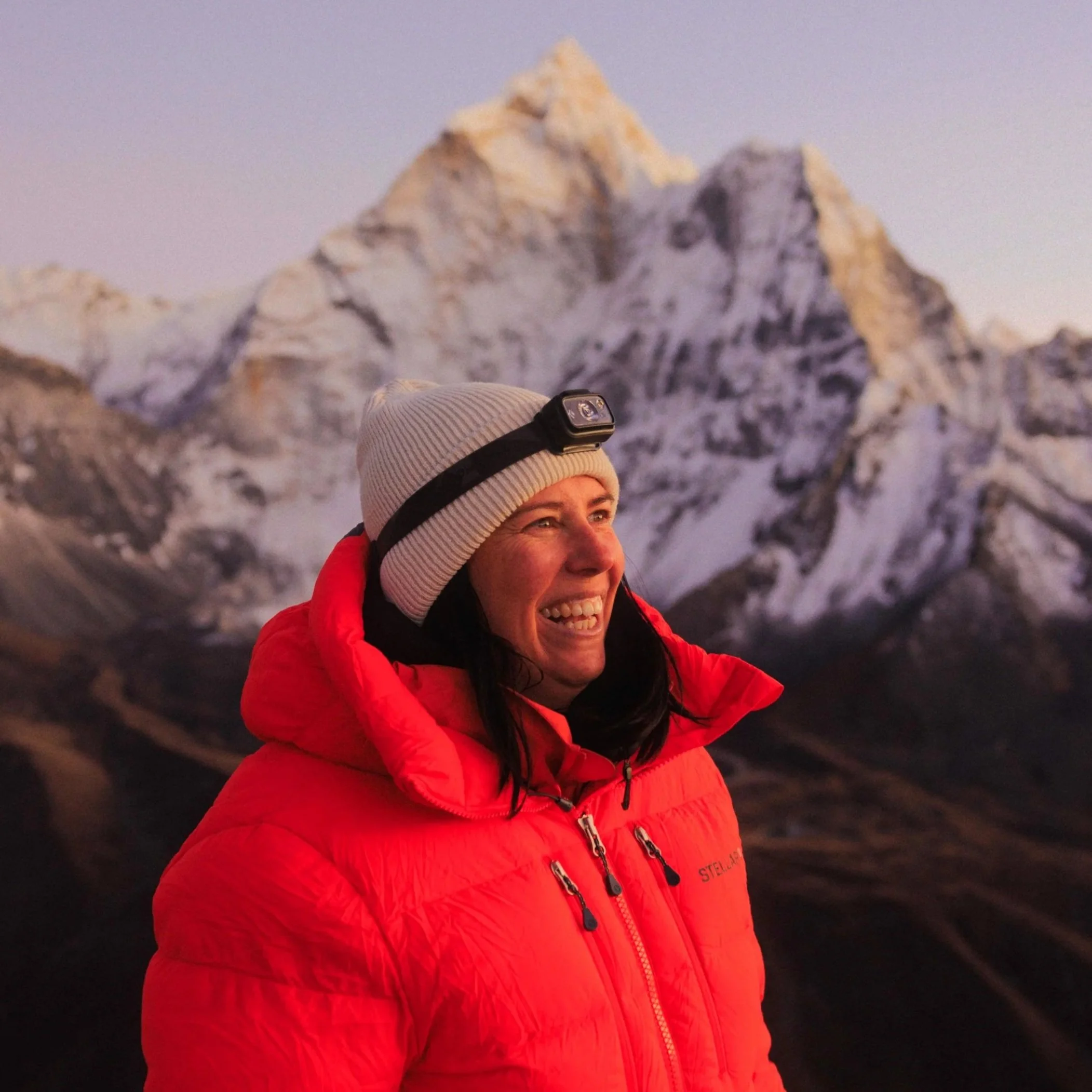 Frau in roter Jacke mit weißen Mütze und Stirnlampe vor schneebedeckten Bergen bei Sonnenuntergang.
