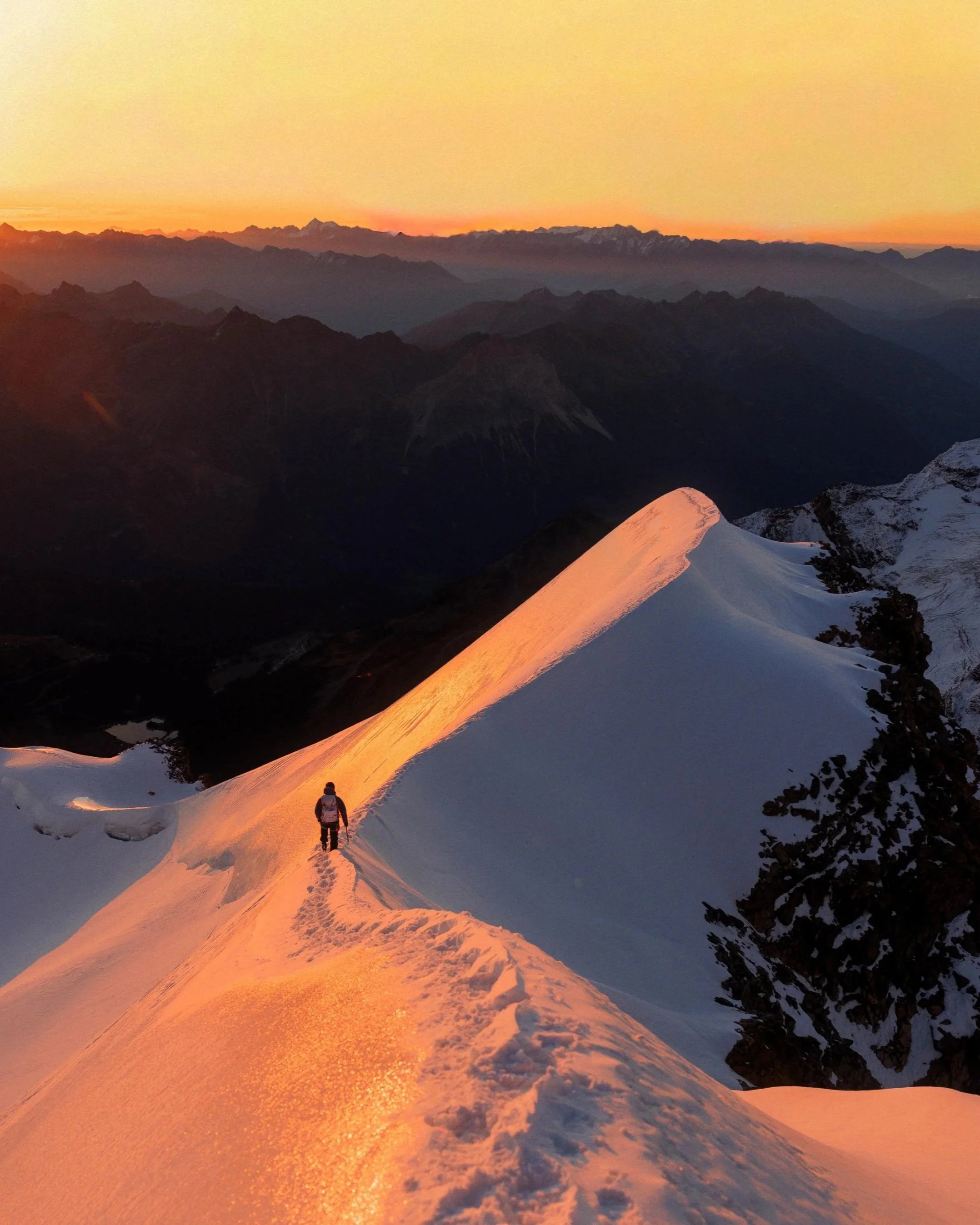 Eine Person wandert auf einem schneebedeckten Berg bei Sonnenuntergang, mit einem farbenprächtigen Himmel im Hintergrund. David Herzig. Fotografie Workshop Nepal. Trekking und Fotografie Reise. Bergfotografie. Sonnenuntergang. Abenteuerfotografie.