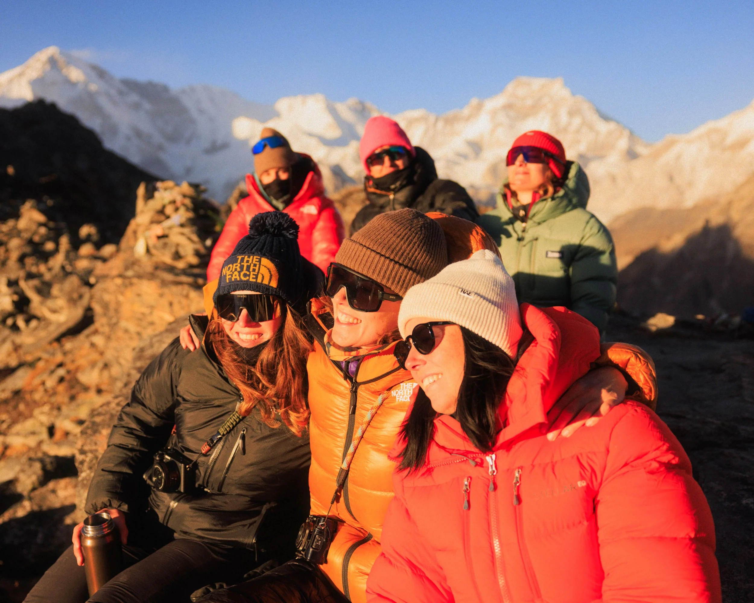 Gruppe von sieben Menschen in Winterkleidung auf einem Berggipfel bei Sonnenuntergang, umgeben von schneebedeckten Bergen.