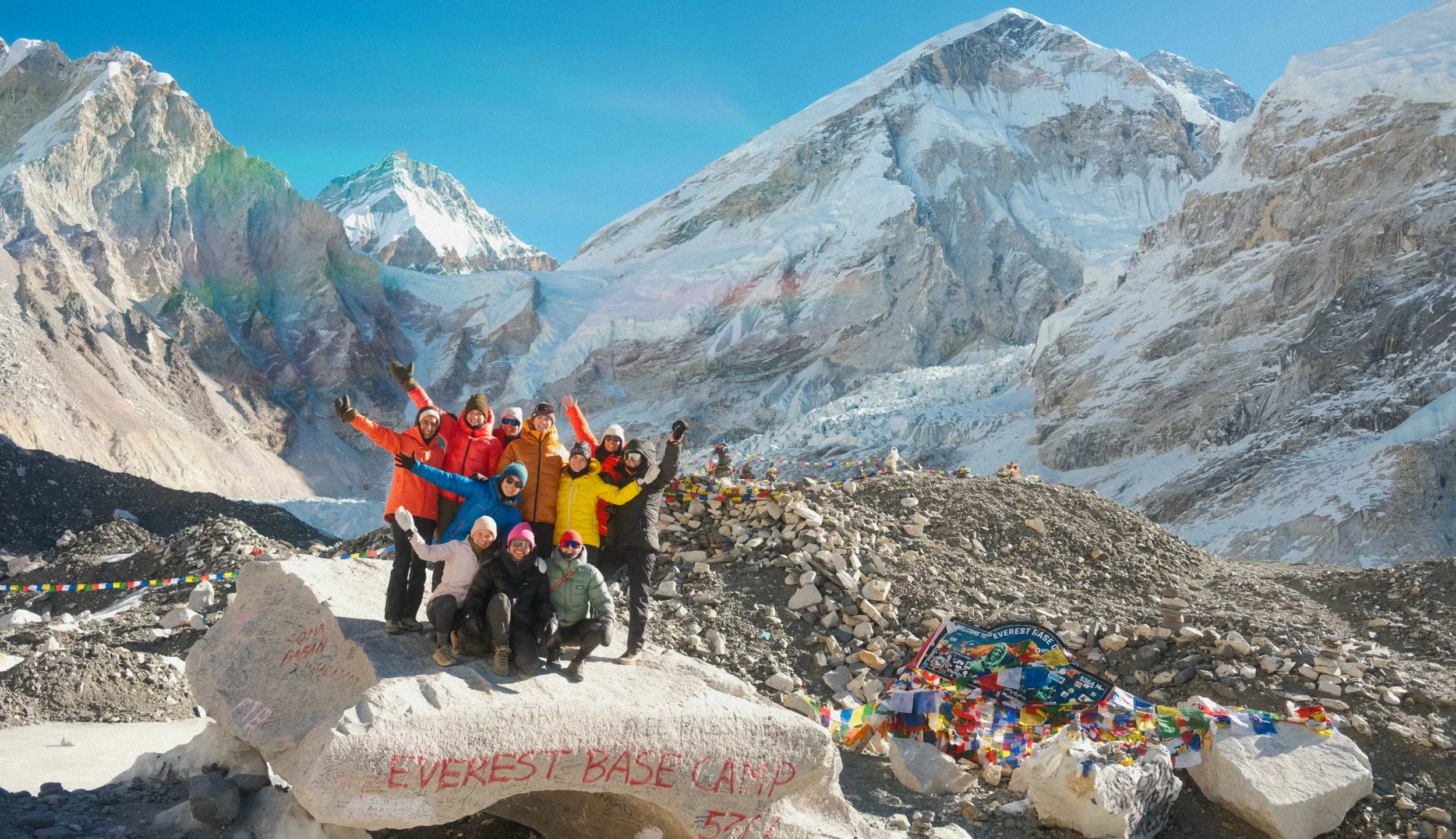 Gruppe von Menschen am Everest Basecamp mit Berggipfeln, Mount Everest und Schneefeldern im Hintergrund, einige tragen bunte Jacken, umgeben von Gebetsflaggen. Individuelle Gruppenreise mit Herz nach Nepal.