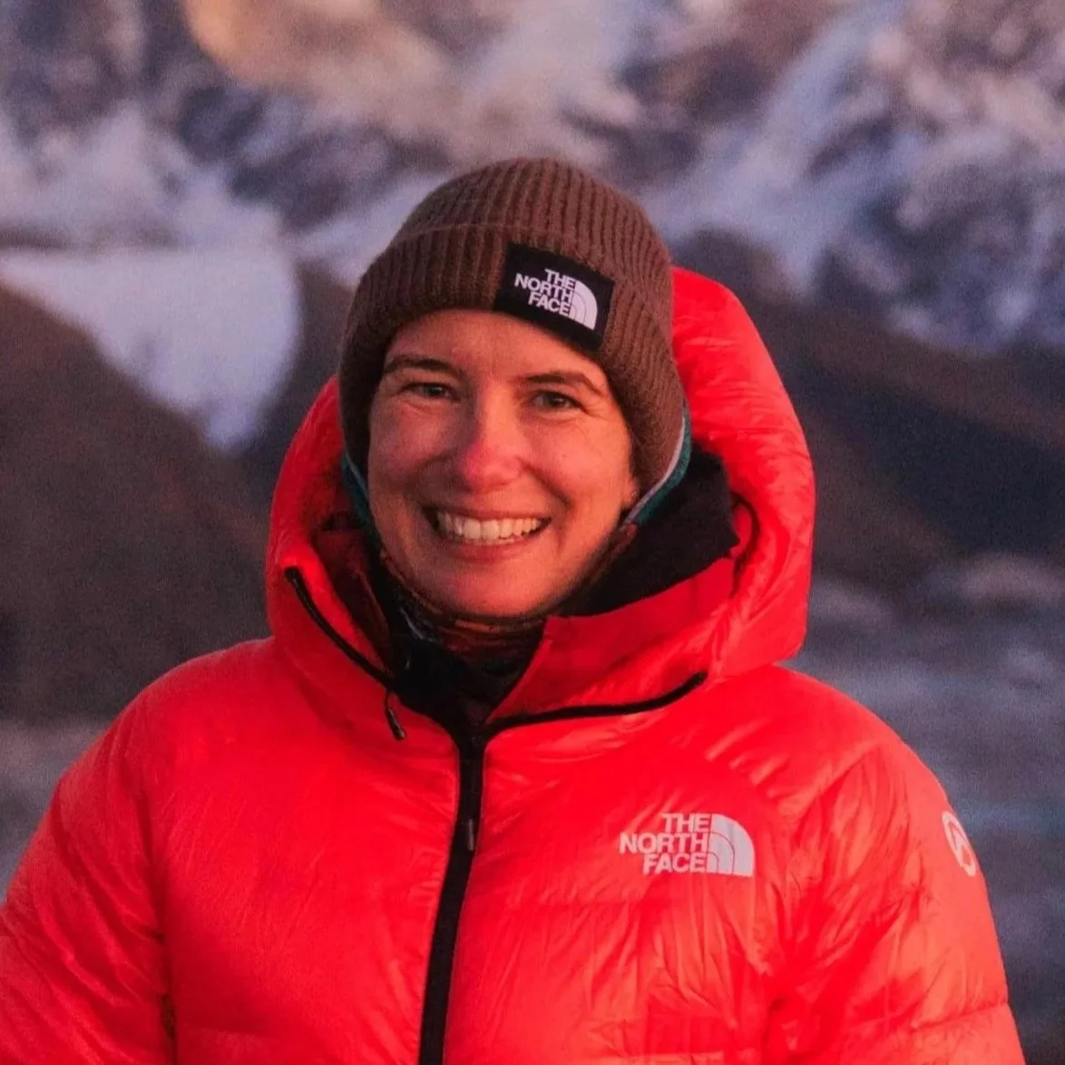 Ein lachender Mann in roter Outdoor-Jacke und brauner Mütze vor einer verschneiten Berglandschaft.