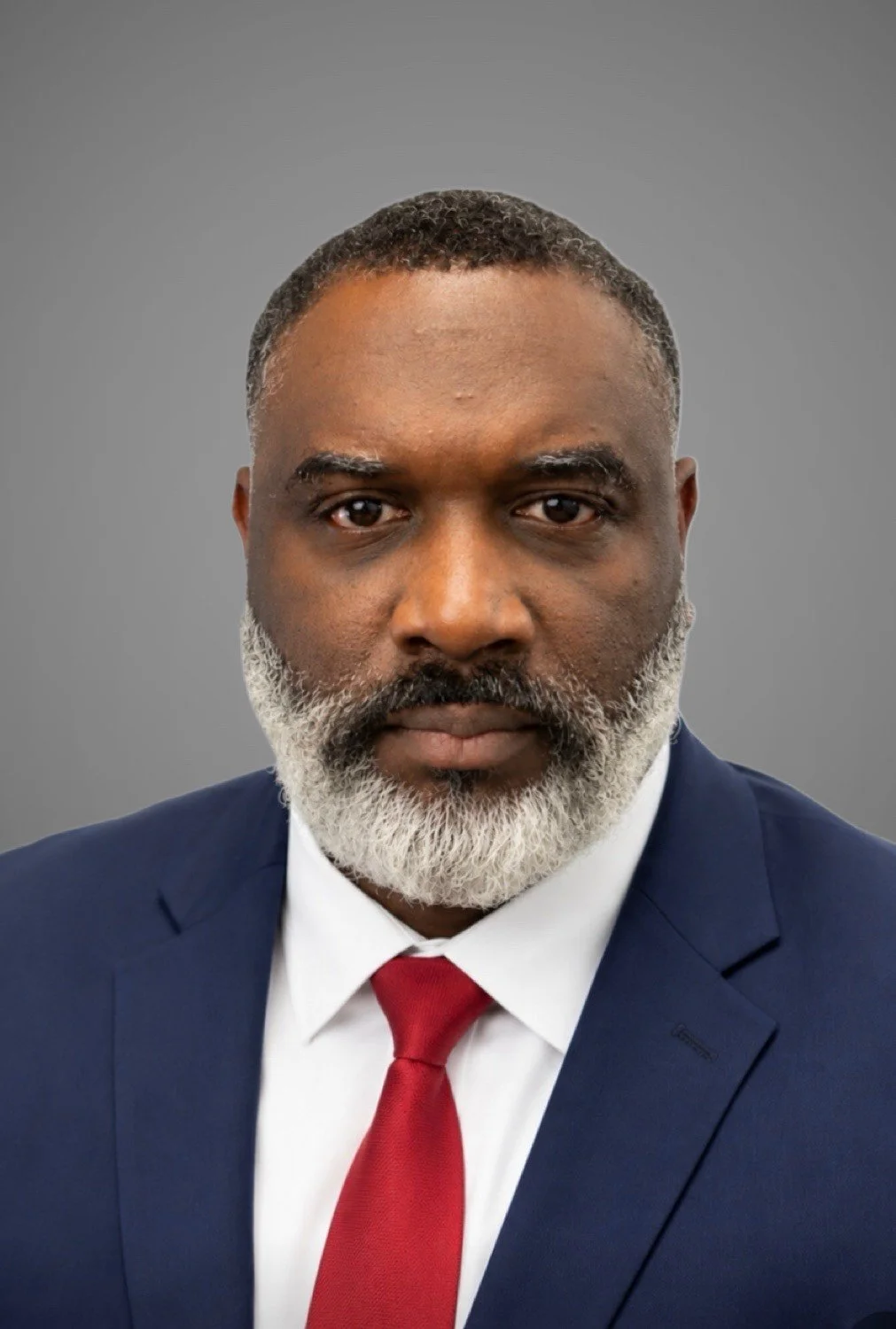 A professional portrait of an African American man with a gray beard, wearing a navy blue suit, white shirt, and red tie, against a plain gray background.