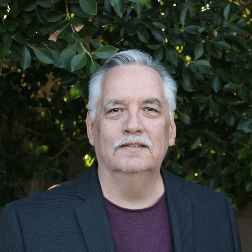 Portrait of a middle-aged man with gray hair and a mustache, wearing a dark blazer and maroon shirt, standing outdoors with green leafy bushes in the background.