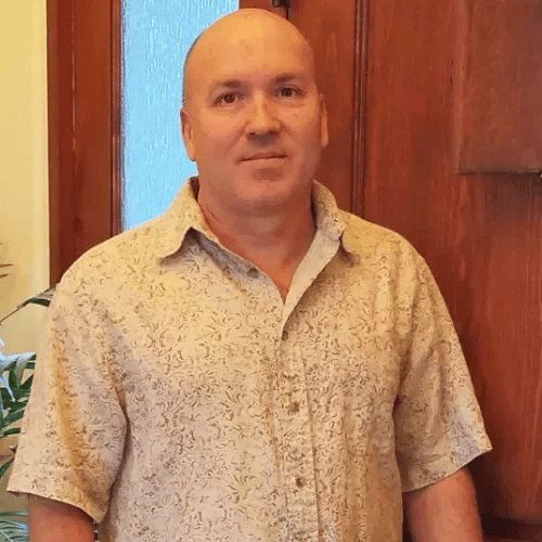 A bald man in a short-sleeve, patterned beige shirt standing indoors in front of wooden paneling and a window.