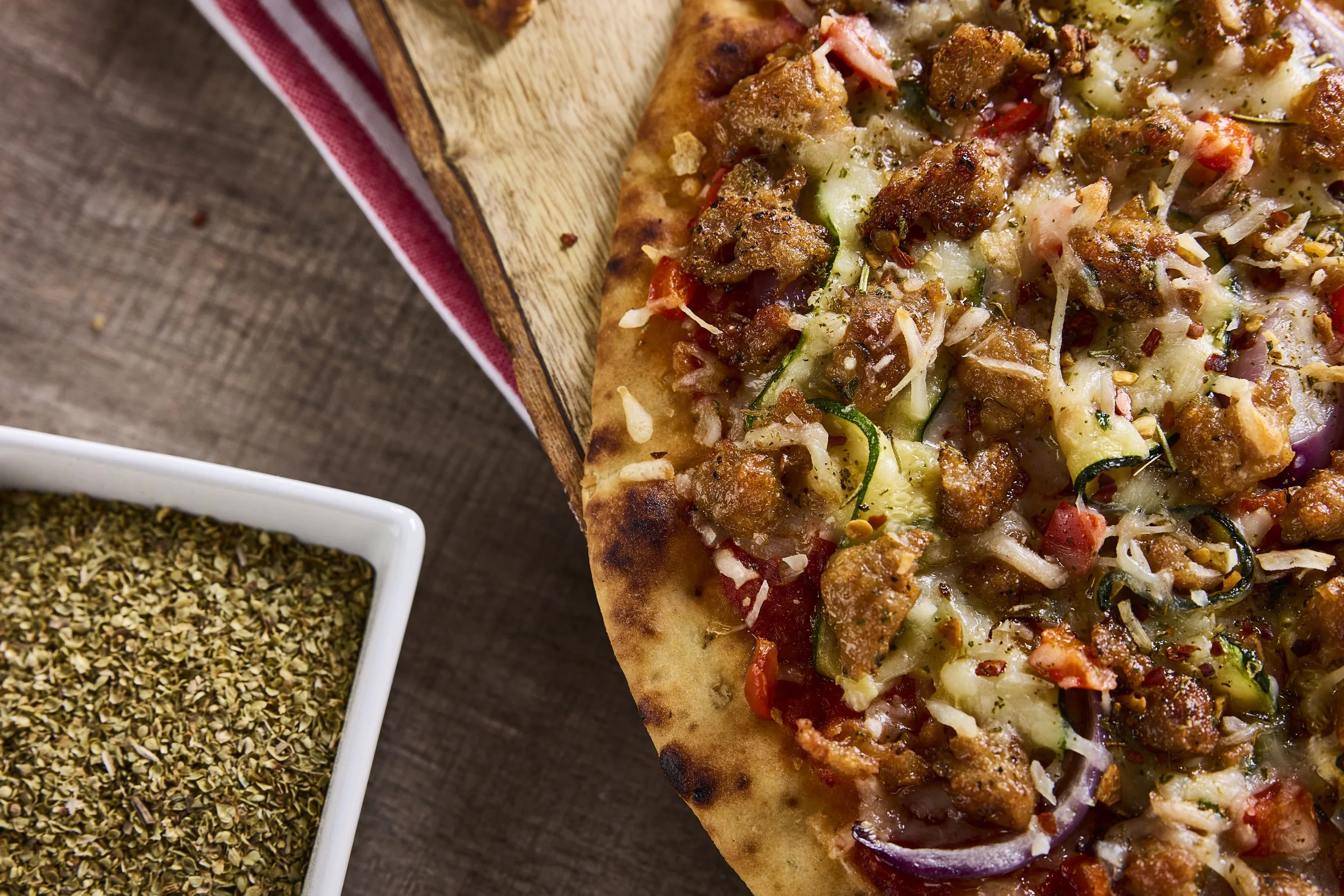 Close-up of a pizza topped with seasoned ground meat, cheese, tomato, onion, and zucchini, on a wooden surface.