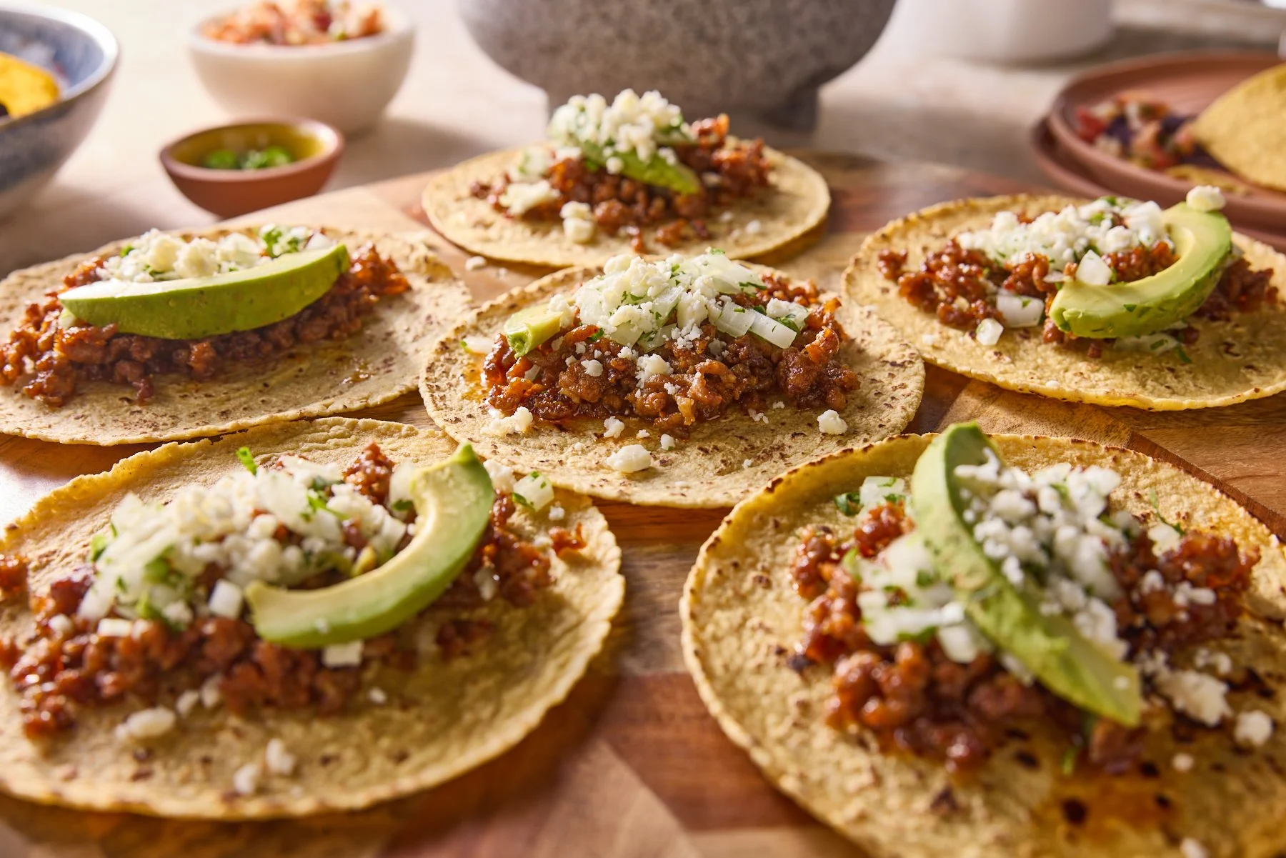 Five tacos on a wooden surface topped with ground meat, shredded cheese, and slices of avocado.