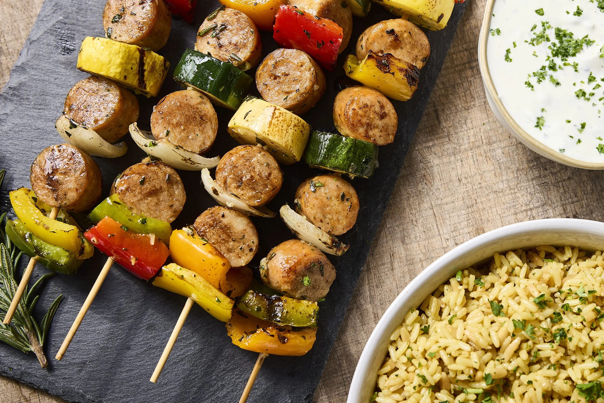 Grilled sausage and vegetable skewers with bowls of rice and creamy herb dressing on a wooden table.