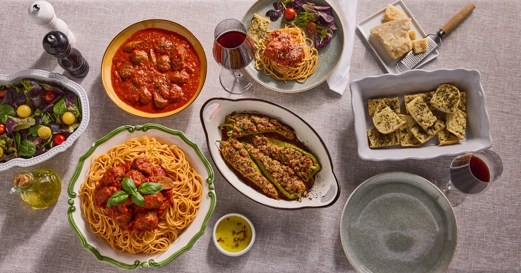 A table set with Italian dishes including spaghetti with meatballs and basil, stuffed green peppers, a vegetable salad, a bowl of spinach and ricotta gnocchi, a bowl of meatballs in tomato sauce, and a side of bread and cheese. There are also glasses