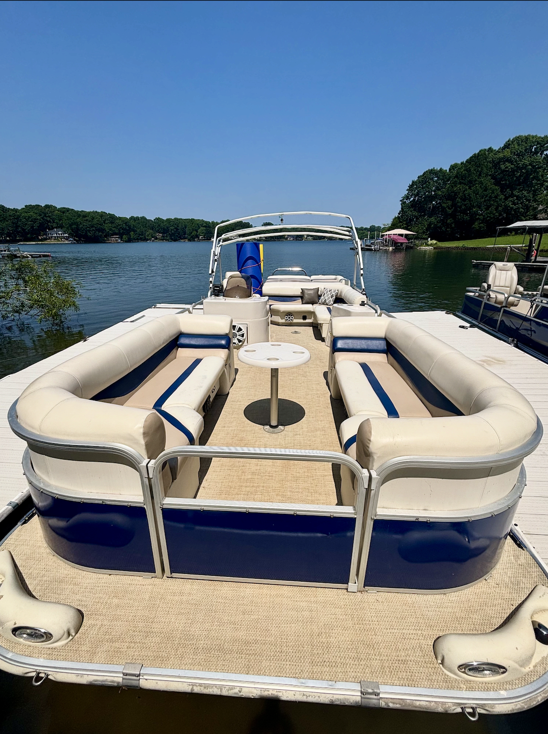 A pontoon boat docked on Lake Norman with beige and blue cushioned seating, a small table, and a clear blue sky.
