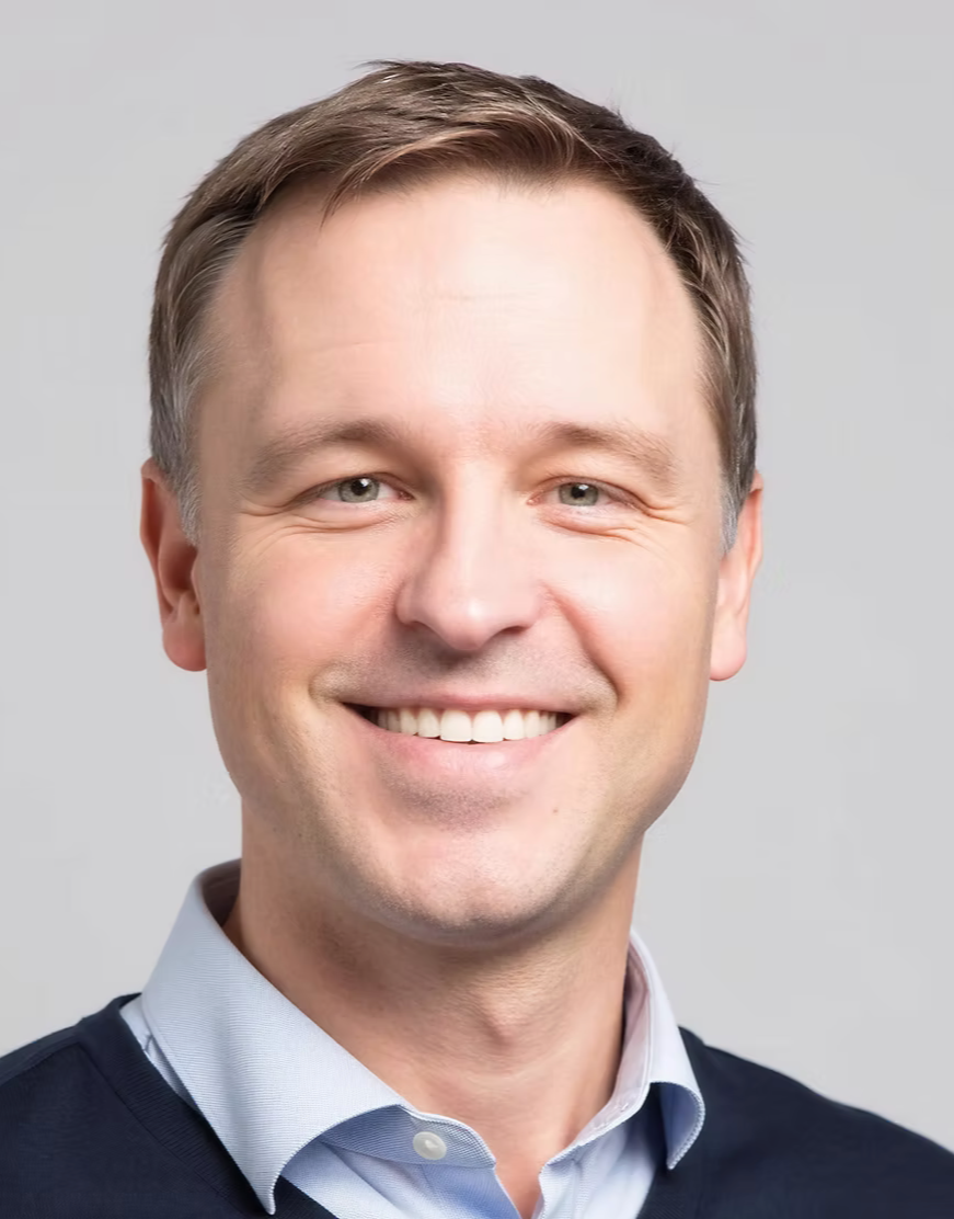 A smiling man with short light brown hair wearing a light blue dress shirt and a dark sweater against a light gray background.
