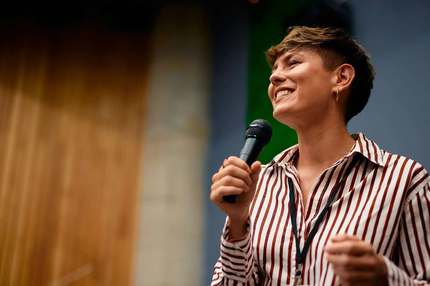 A woman with short hair, wearing a striped shirt and hoop earrings, holding a microphone and smiling during a speech or presentation.