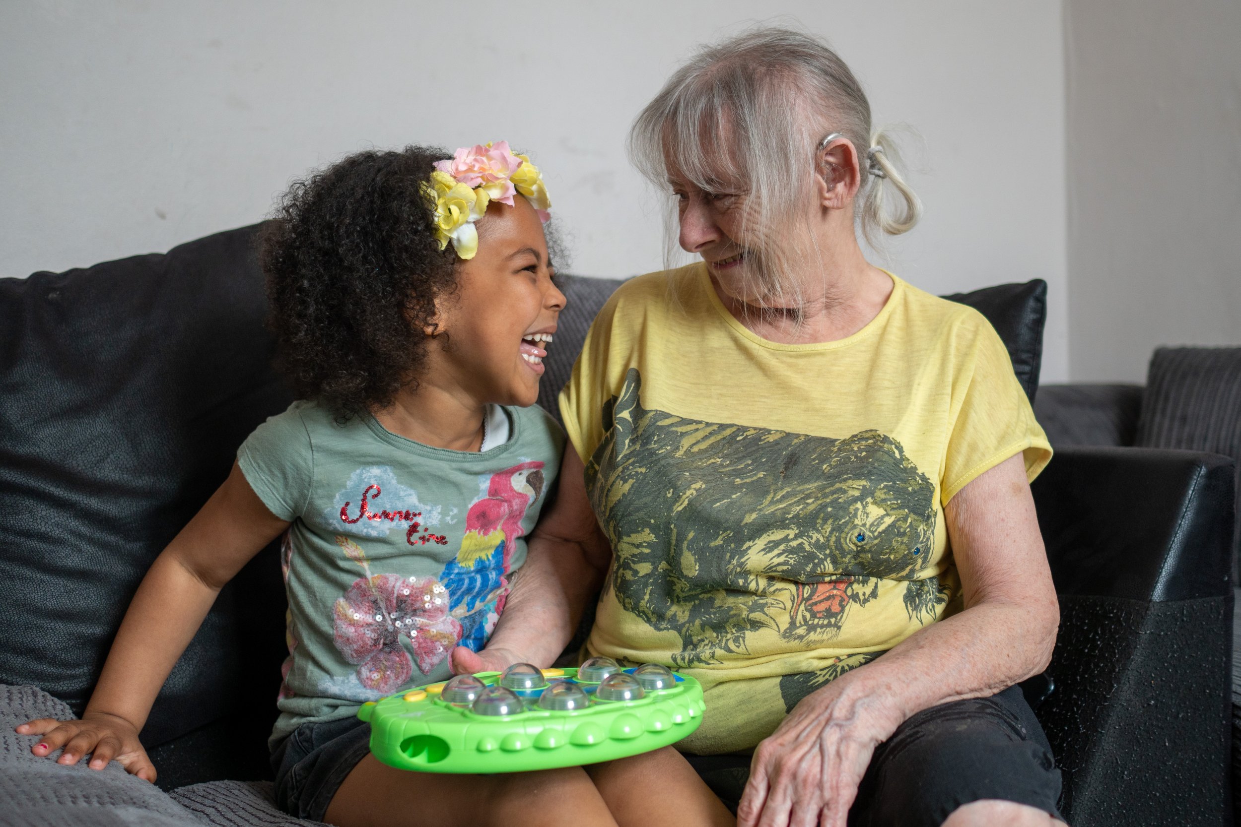 An elderly woman and a young girl sitting on a black sofa, smiling and laughing at each other.