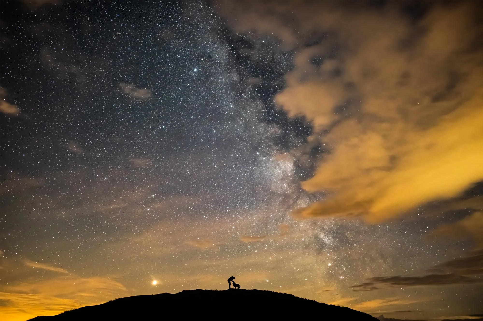 Silhouette of a person on a hill under a star-filled night sky with the Milky Way galaxy and some clouds.