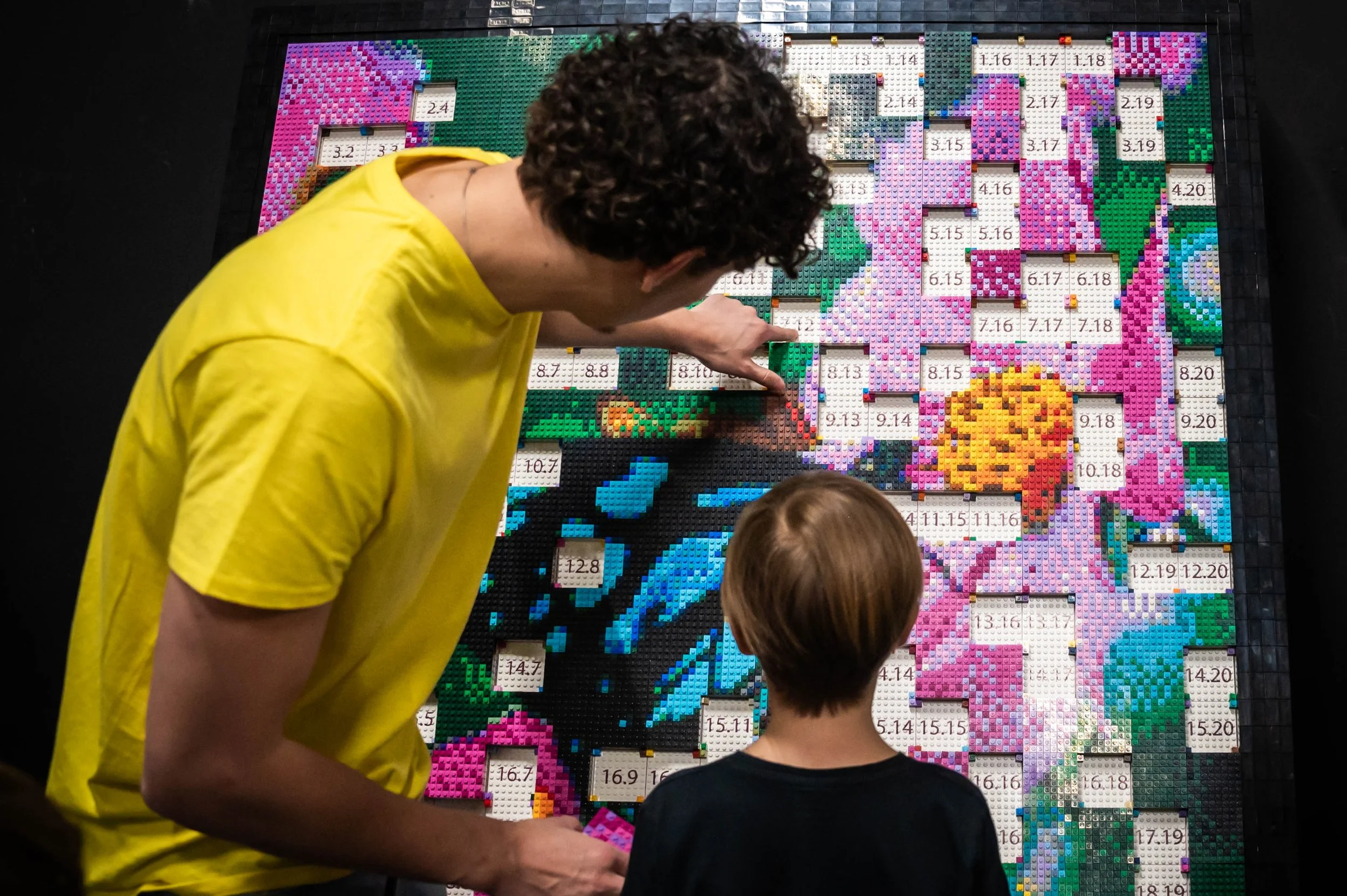 Zwei Personen, ein Erwachsener in gelbem T-Shirt und ein Kind, betrachten ein großes Bild aus LEGO-Steinen, das eine bunte Blumenmuster zeigt.