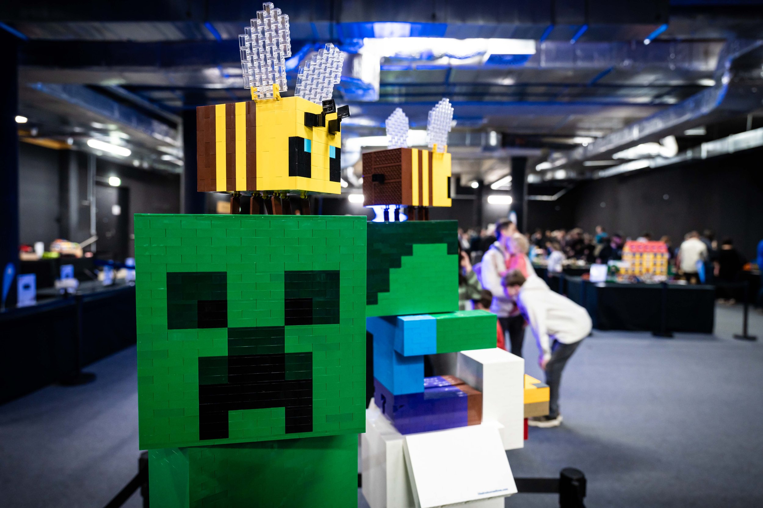 Lego-Skulptur einer grünen Creeper-Figur aus dem Spiel Minecraft, mit einer gelben Biene auf dem Kopf, in einer Ausstellungshalle mit mehreren Besuchern im Hintergrund.