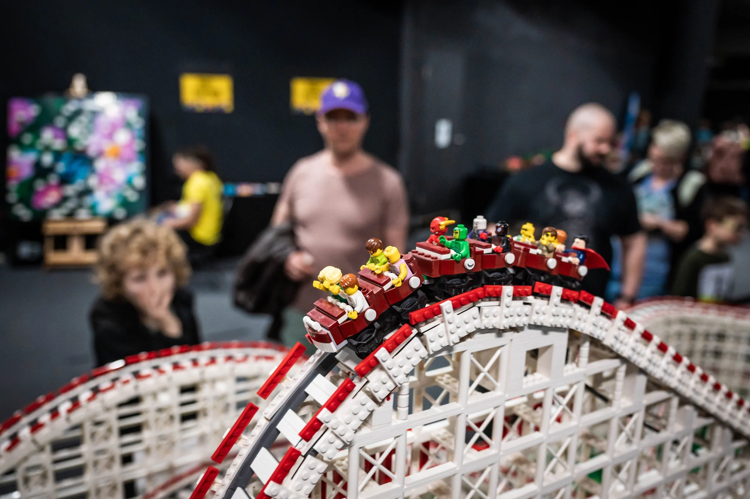 Eine LEGO-Bahn mit Minifiguren, die eine Achterbahn darstellen, im Vordergrund. Im Hintergrund sind mehrere Menschen, die die Ausstellung betrachten.